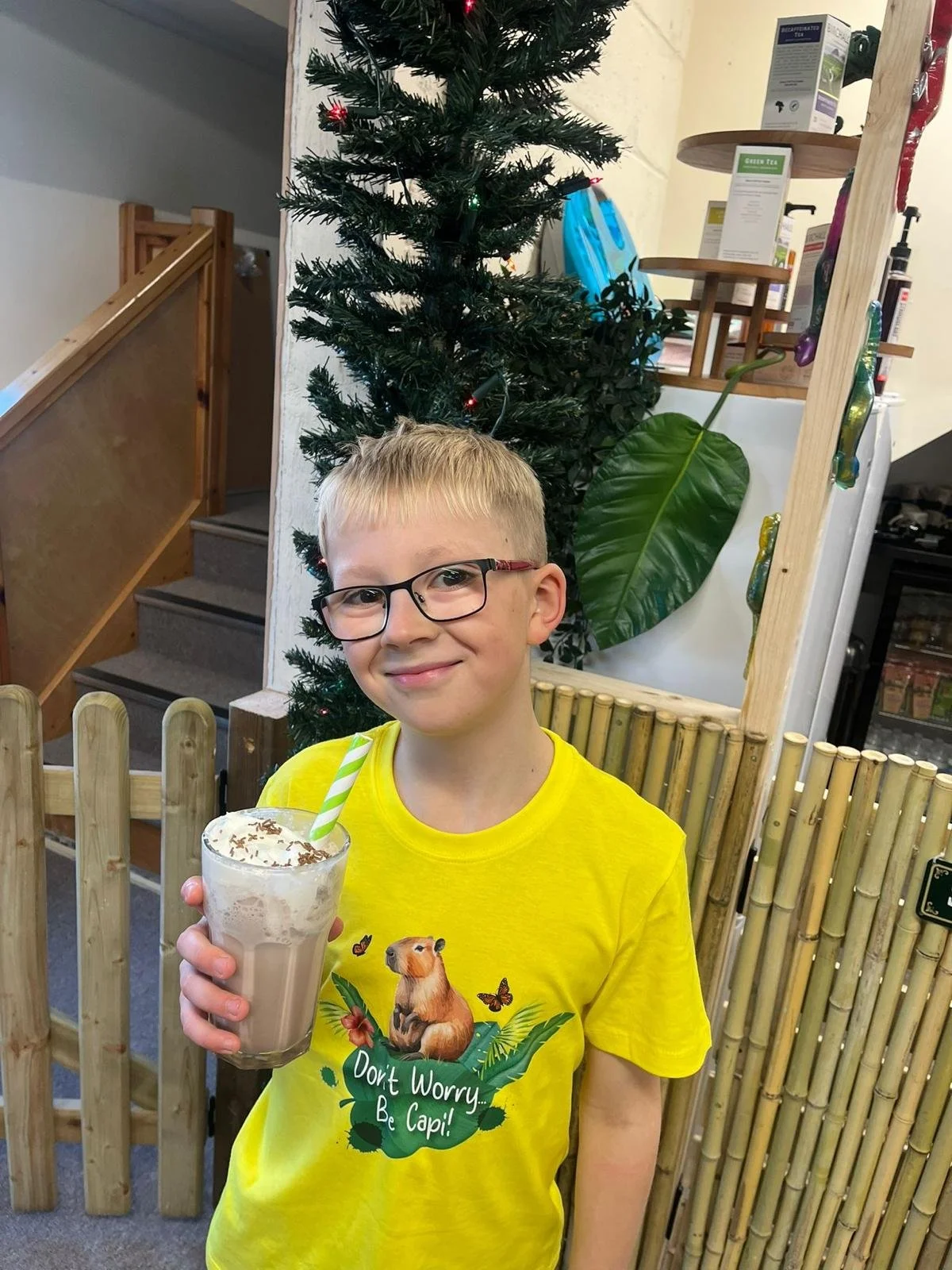 A young boy with glasses and a yellow t-shirt holding a milkshake with whipped cream and a green striped straw, standing in front of a Christmas tree and bamboo fence inside a store.