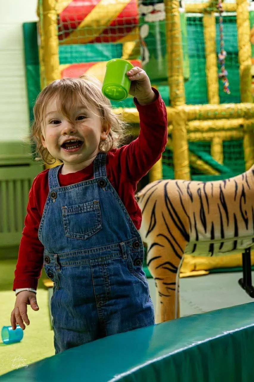A young child with curly blonde hair, smiling and holding a green cup up in a play area with jungle-themed decorations, including a fabric tiger and a colorful jungle gym in the background.