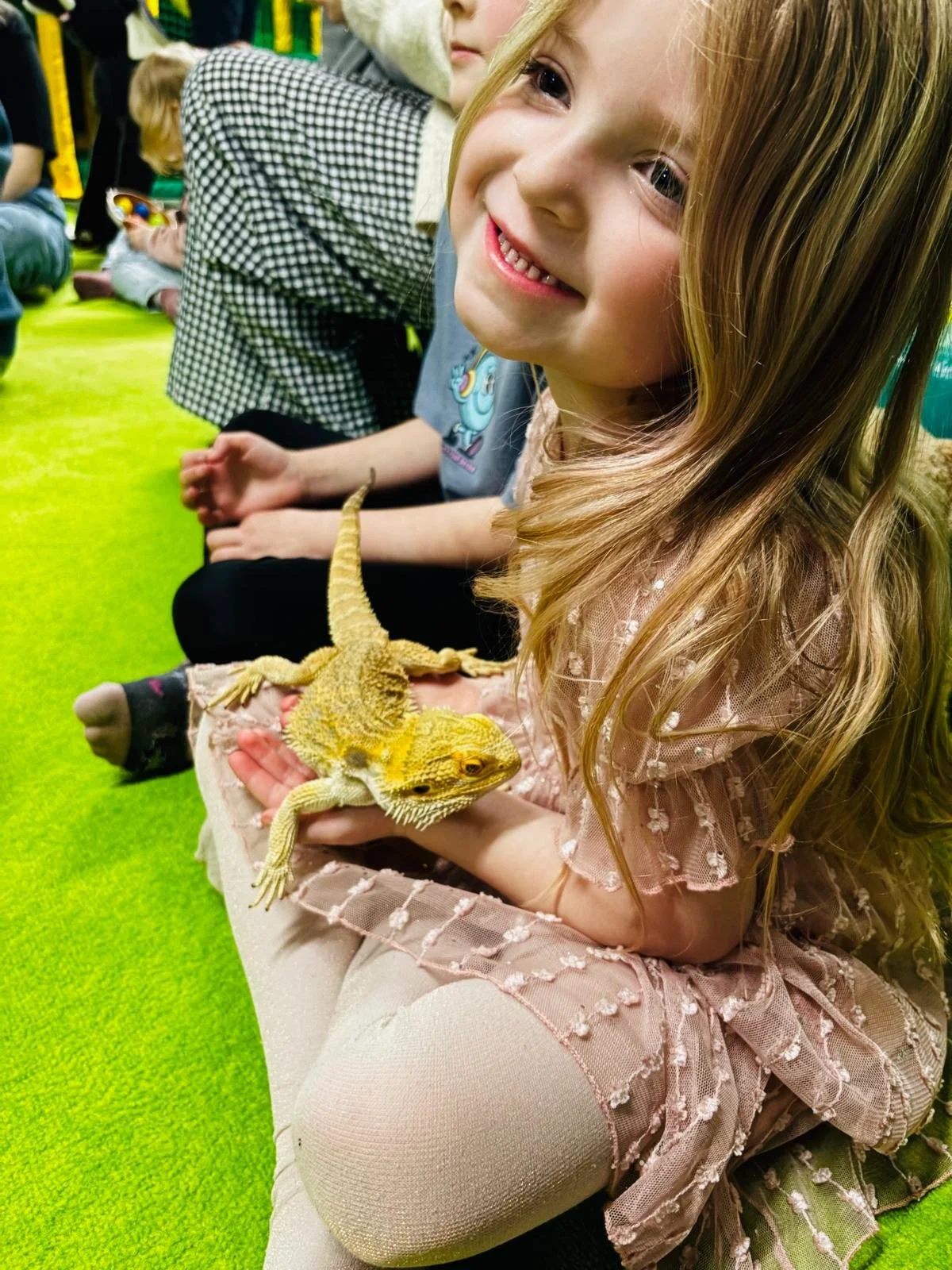 A young girl with long blonde hair smiling while holding a yellow bearded dragon. She is sitting on a green carpet with other children in the background, some sitting and some holding toys.