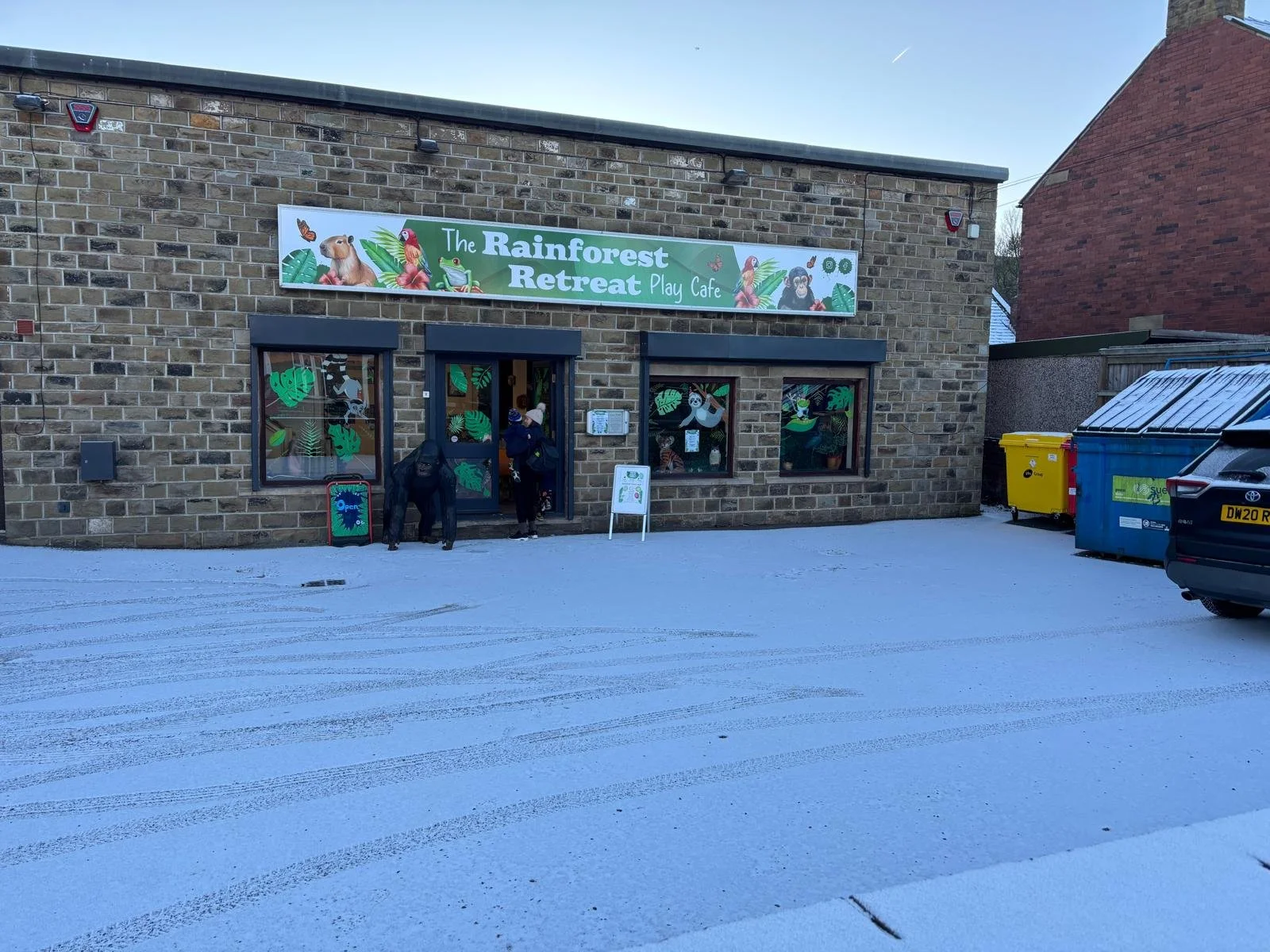 Front view of a building with a sign that reads 'The Rainforest Retreat Play Cafe' and illustrations of rainforest animals, with snow on the ground and a car parked nearby.