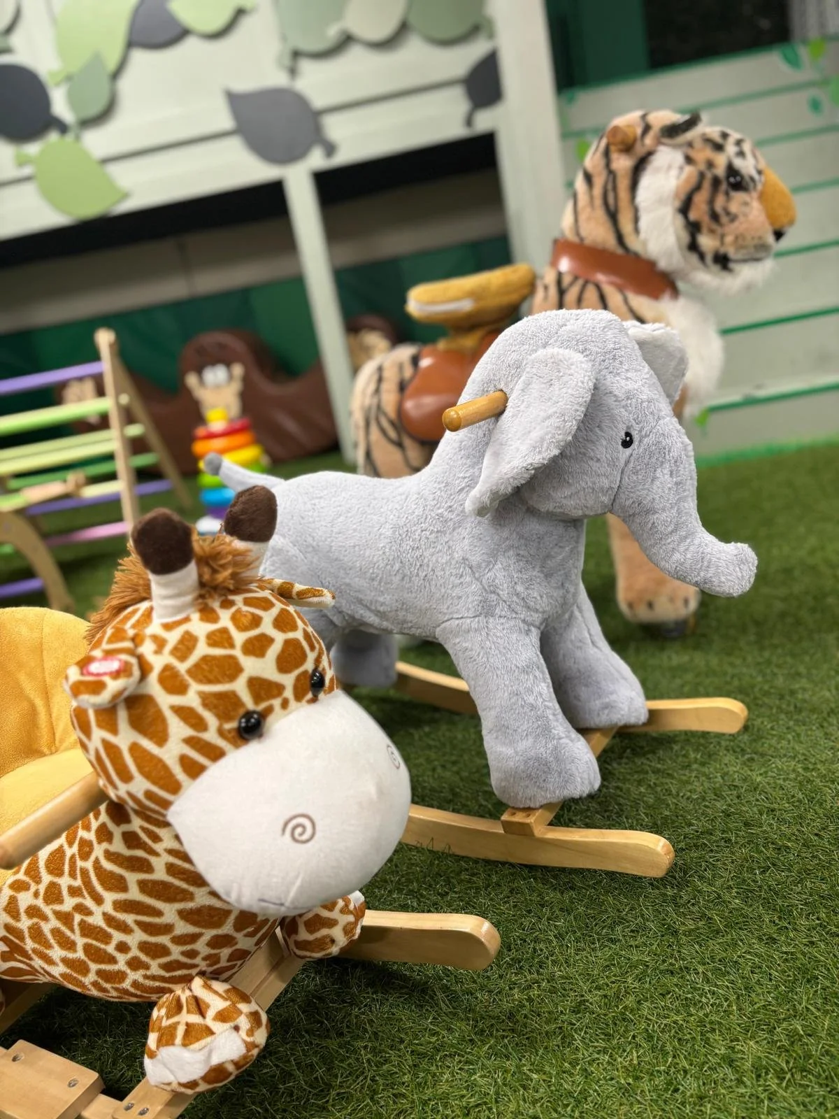 A display of plush animal toys, including a giraffe, an elephant, and a tiger, in a setting with green grass flooring and decorative background elements.