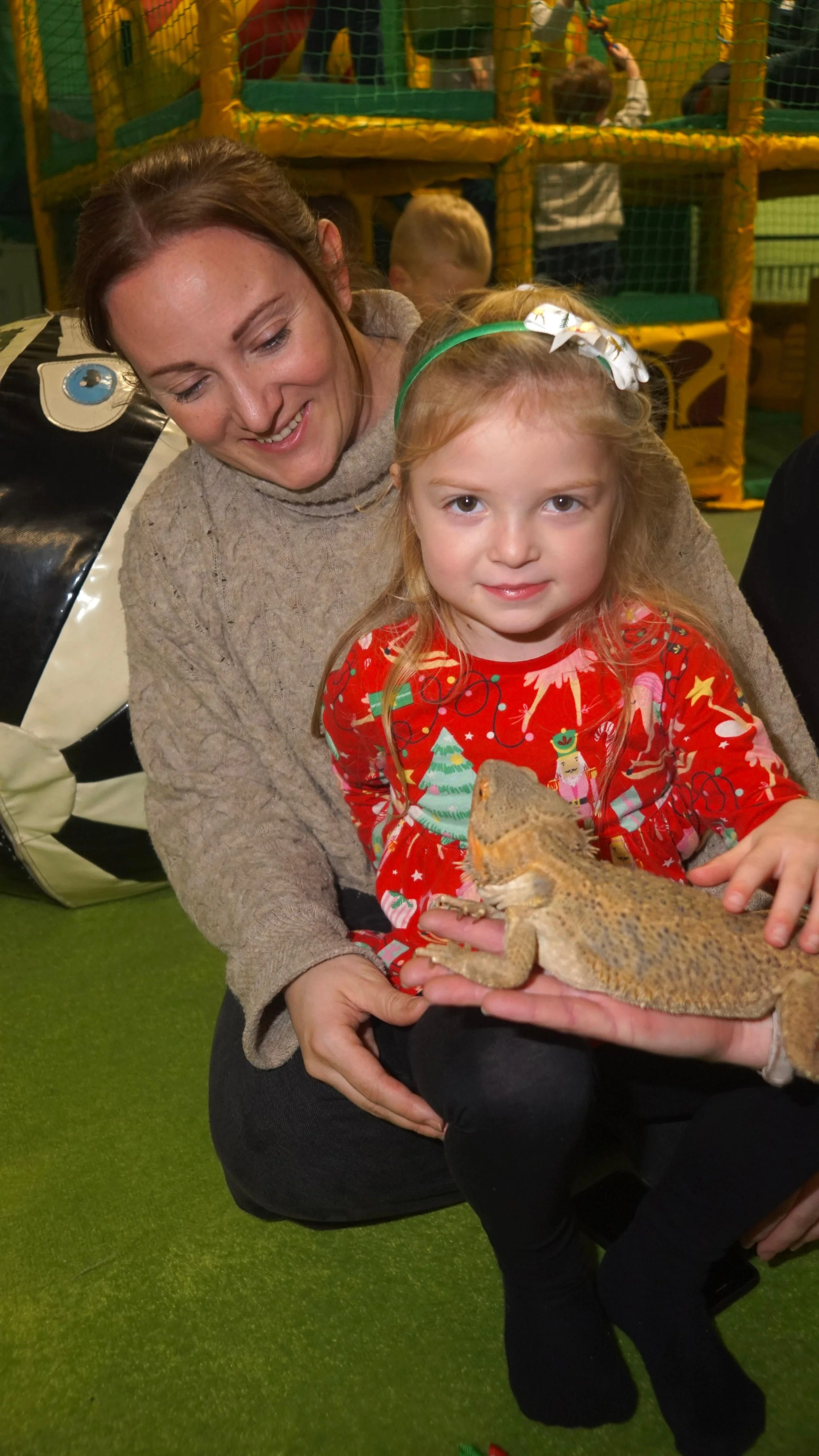 A woman and a young girl holding a bearded dragon lizard, with the woman smiling and the girl looking at the camera, in an indoor play area.