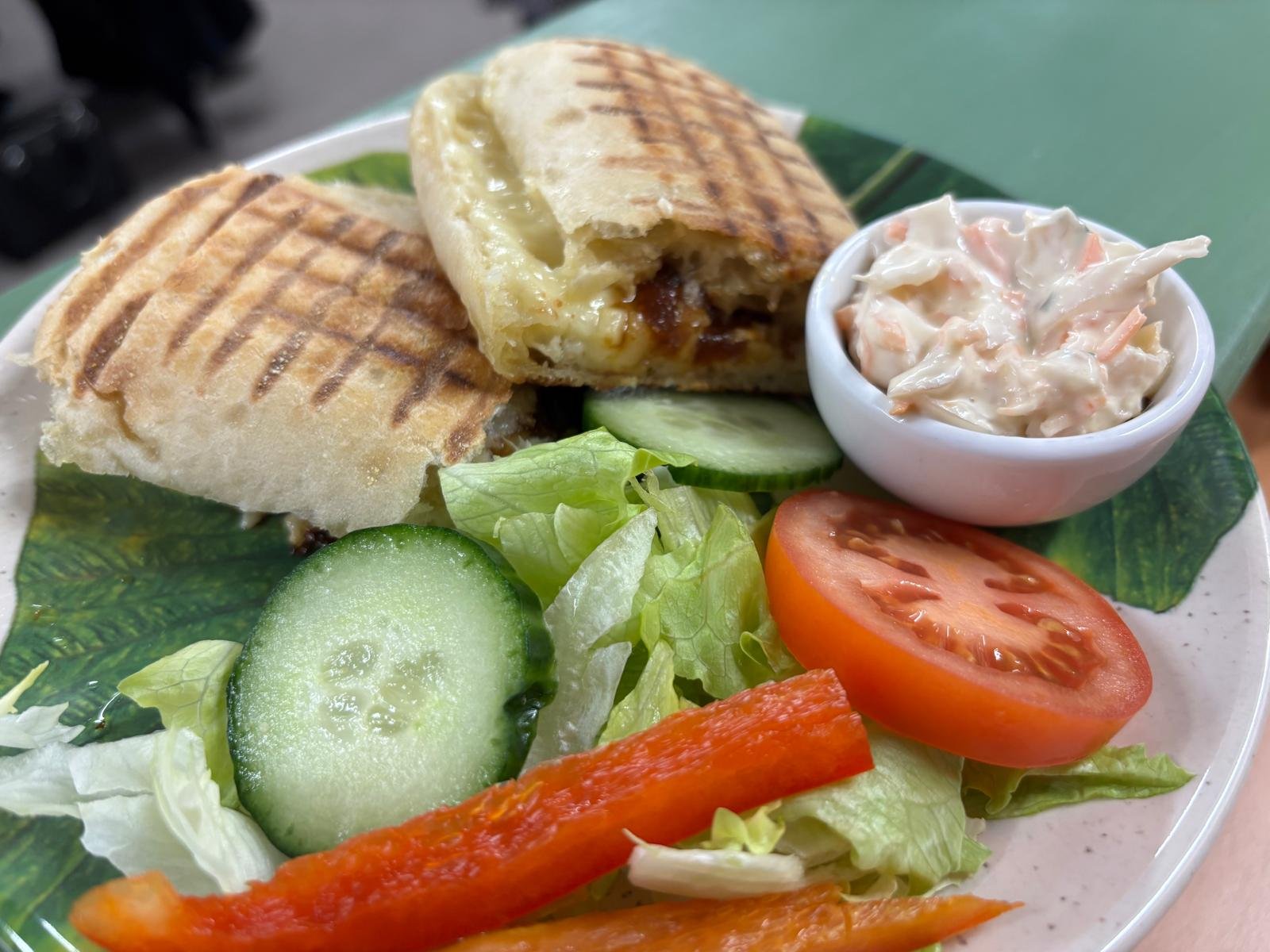 A plate with a grilled sandwich, a small bowl of coleslaw, sliced tomato, cucumber, lettuce, and red bell pepper.