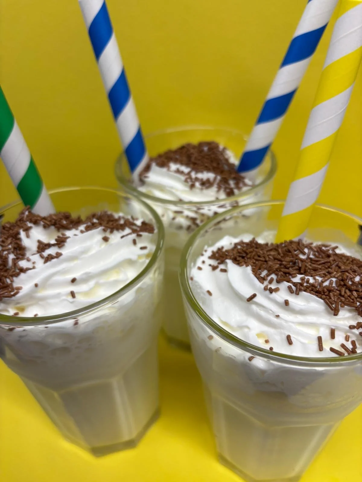 Three glasses filled with a creamy dessert topped with whipped cream and chocolate sprinkles, with colorful striped straws, against a yellow background.