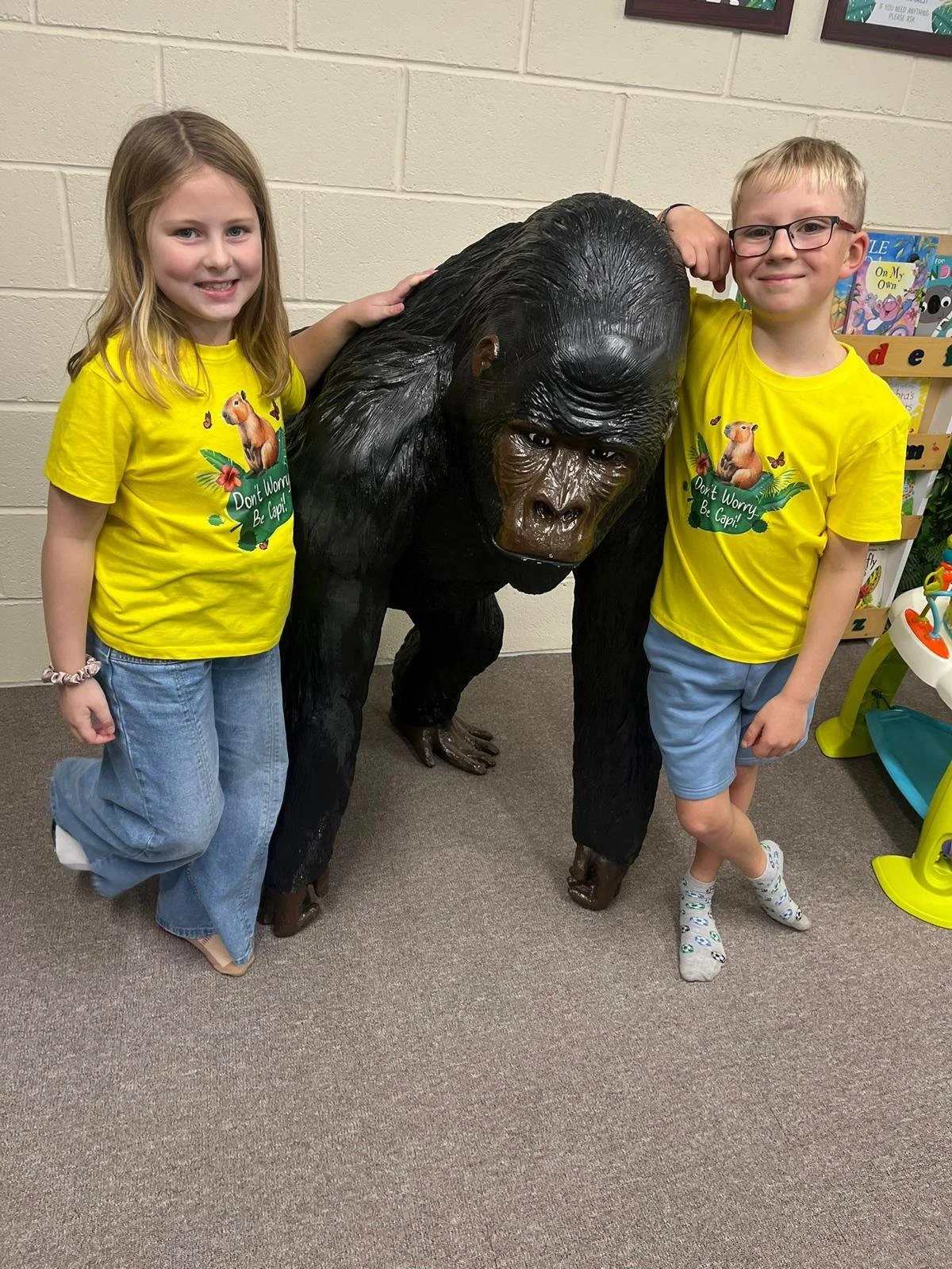 Two children, a girl and a boy, smiling and posing with a large gorilla statue inside a room with beige brick walls. The children are wearing matching yellow t-shirts with a guinea pig and the words 'Don't Worry Be Capy.' The girl has long blonde hai