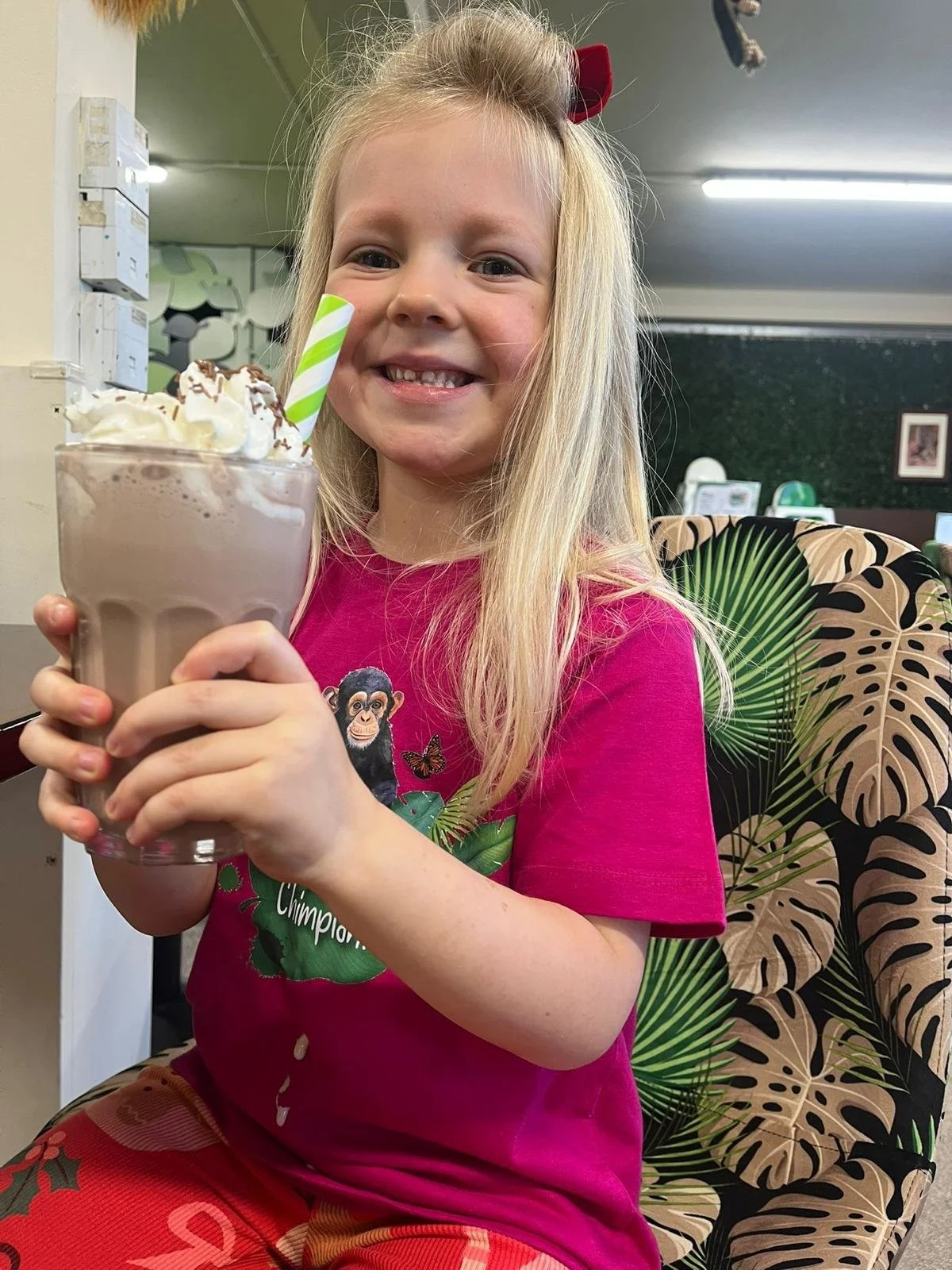 A young girl with long blonde hair, wearing a pink shirt with a monkey and butterfly on it, is sitting in a chair and holding a large glass of chocolate milkshake topped with whipped cream and chocolate shavings. She is smiling at the camera.