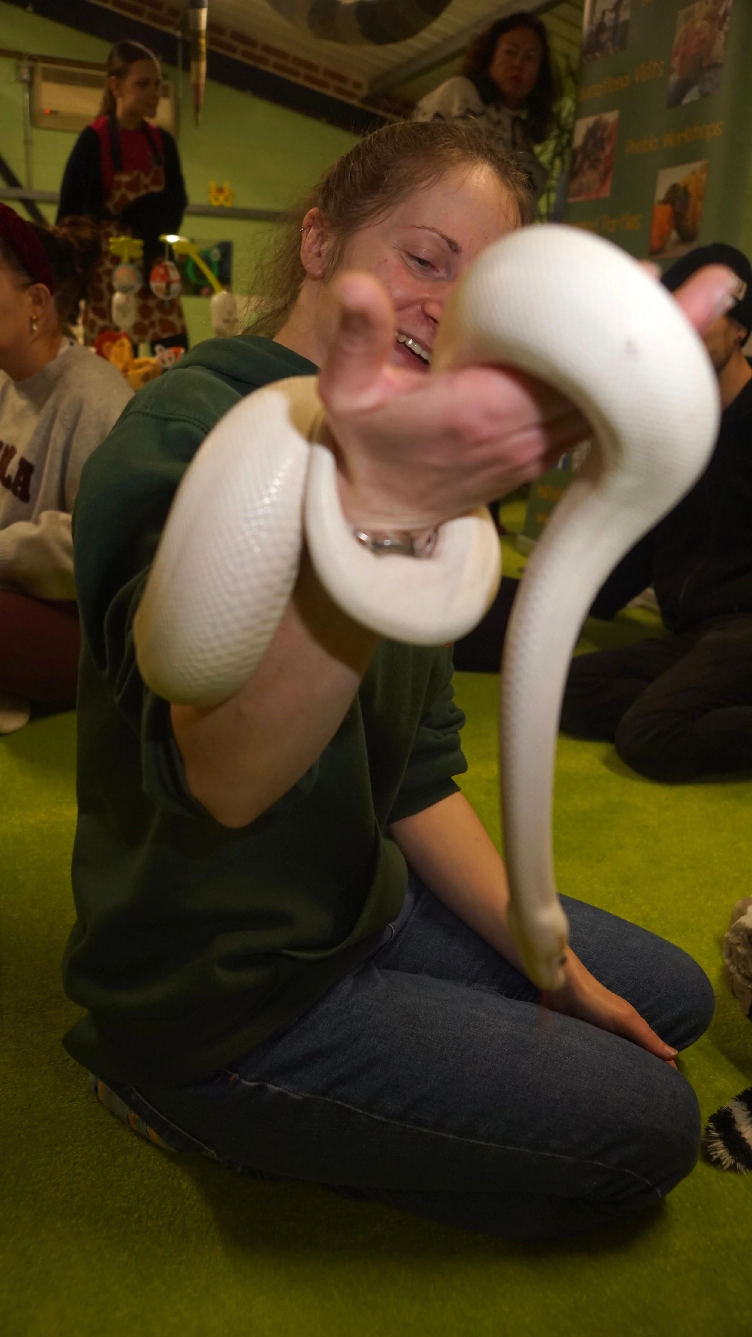 A woman sitting on the green carpet holding a white snake, smiling, surrounded by other people in an indoor environment with colorful decorations.