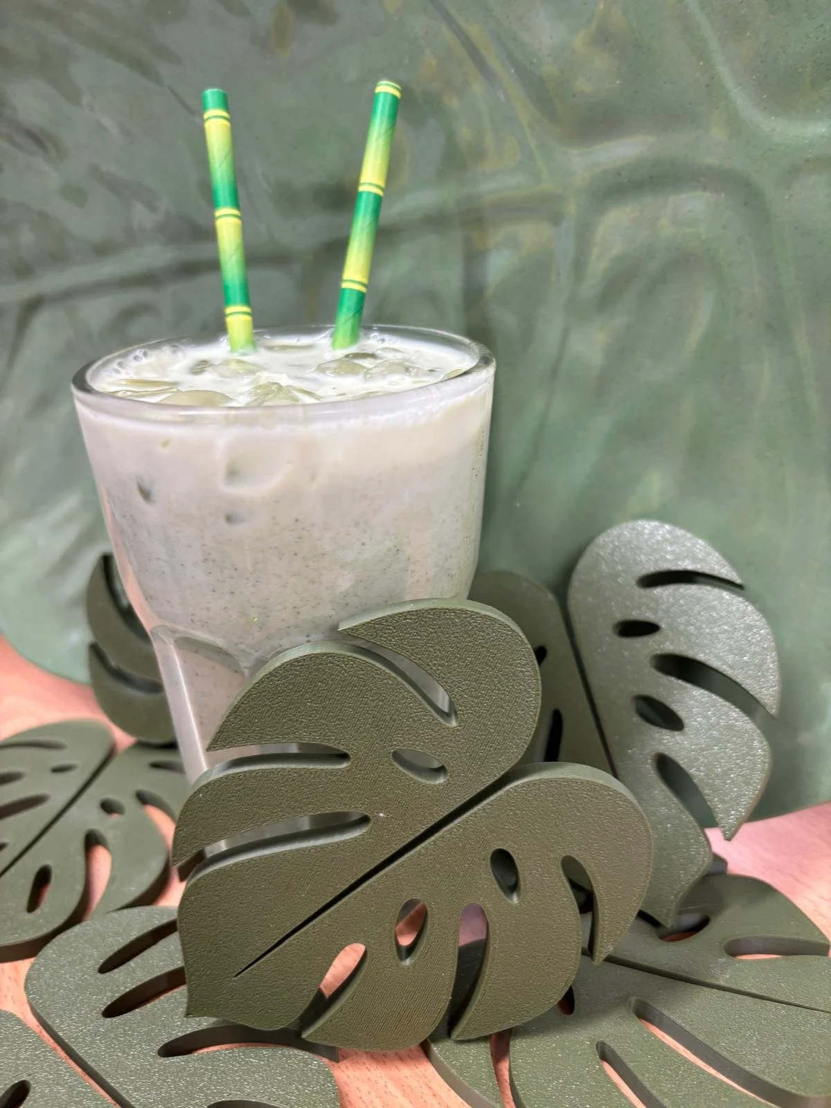 A glass of a creamy, iced beverage with two green-striped straws, surrounded by black decorative monstera leaf-shaped coasters, on a pink surface with a green abstract background.