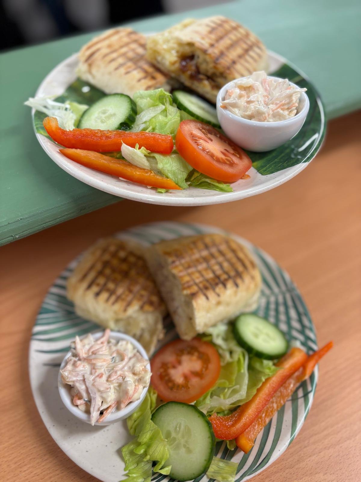 Two plates of grilled sandwiches with salad and coleslaw, one atop a green surface and the other on a wooden table.