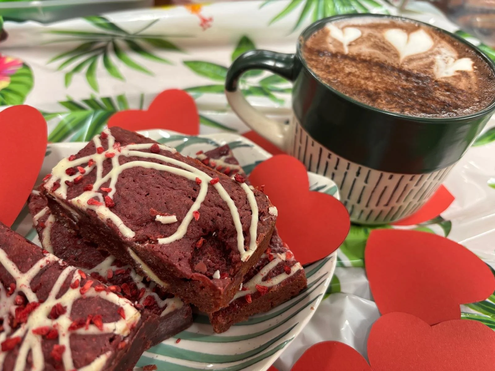 Chocolate brownie topped with white icing and red sprinkles, served with a mug of hot chocolate decorated with white marshmallows and sprinkled cocoa powder, surrounded by red paper hearts on a tropical leaf print tablecloth.