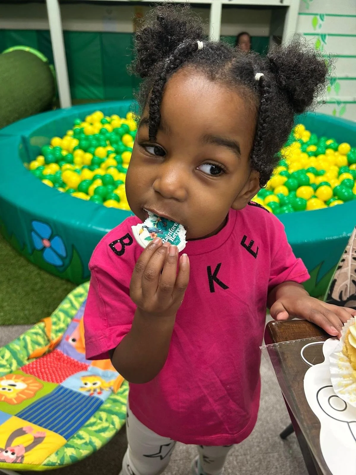 A young girl with braided hair in puffs wearing a pink shirt eating cake in front of a ball pit filled with yellow and green balls.