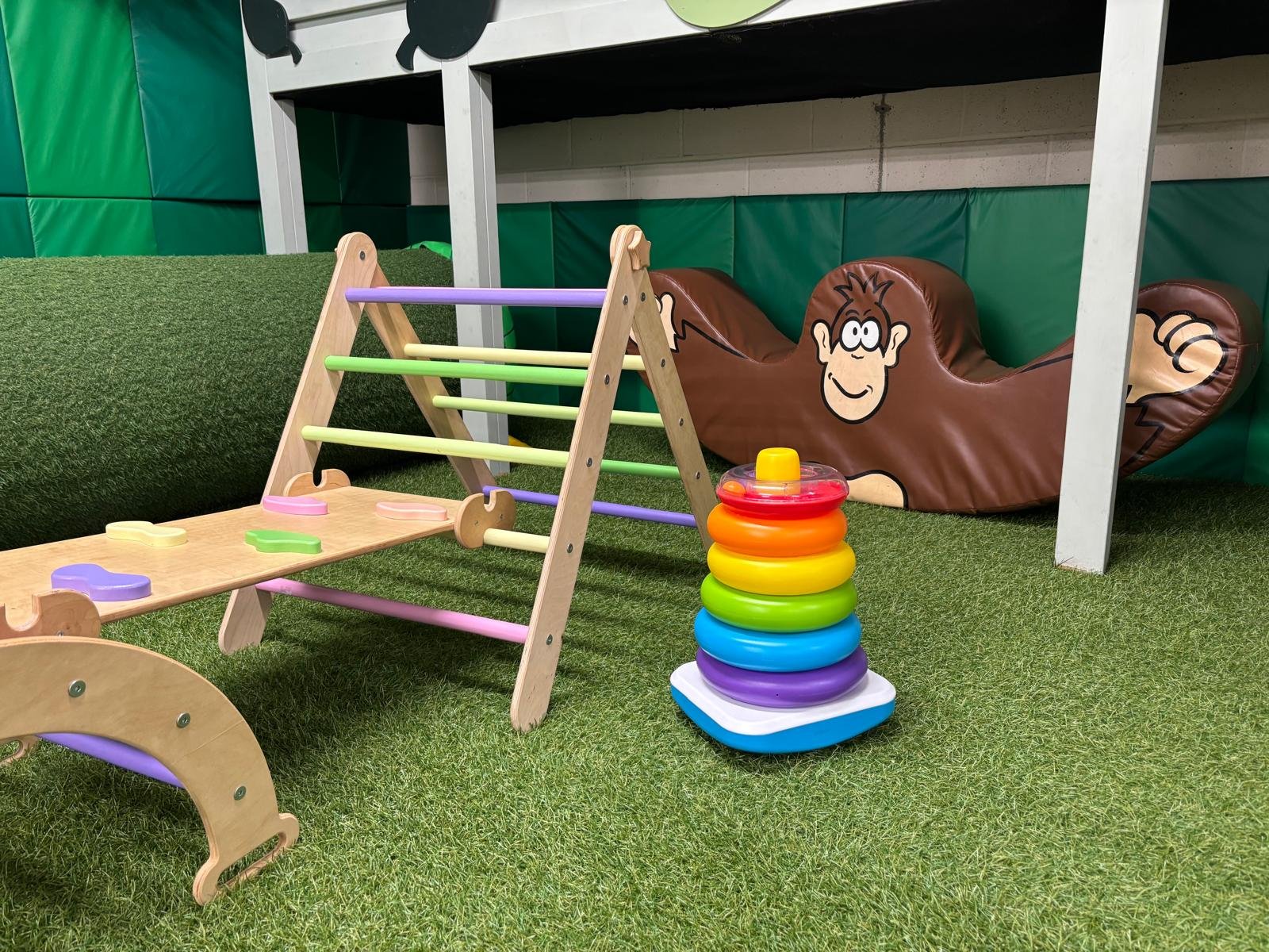 Colorful children’s playground with a wooden climbing structure, a rainbow-colored stacking toy, and an animal-shaped cushion with a cartoon face, all on artificial grass.