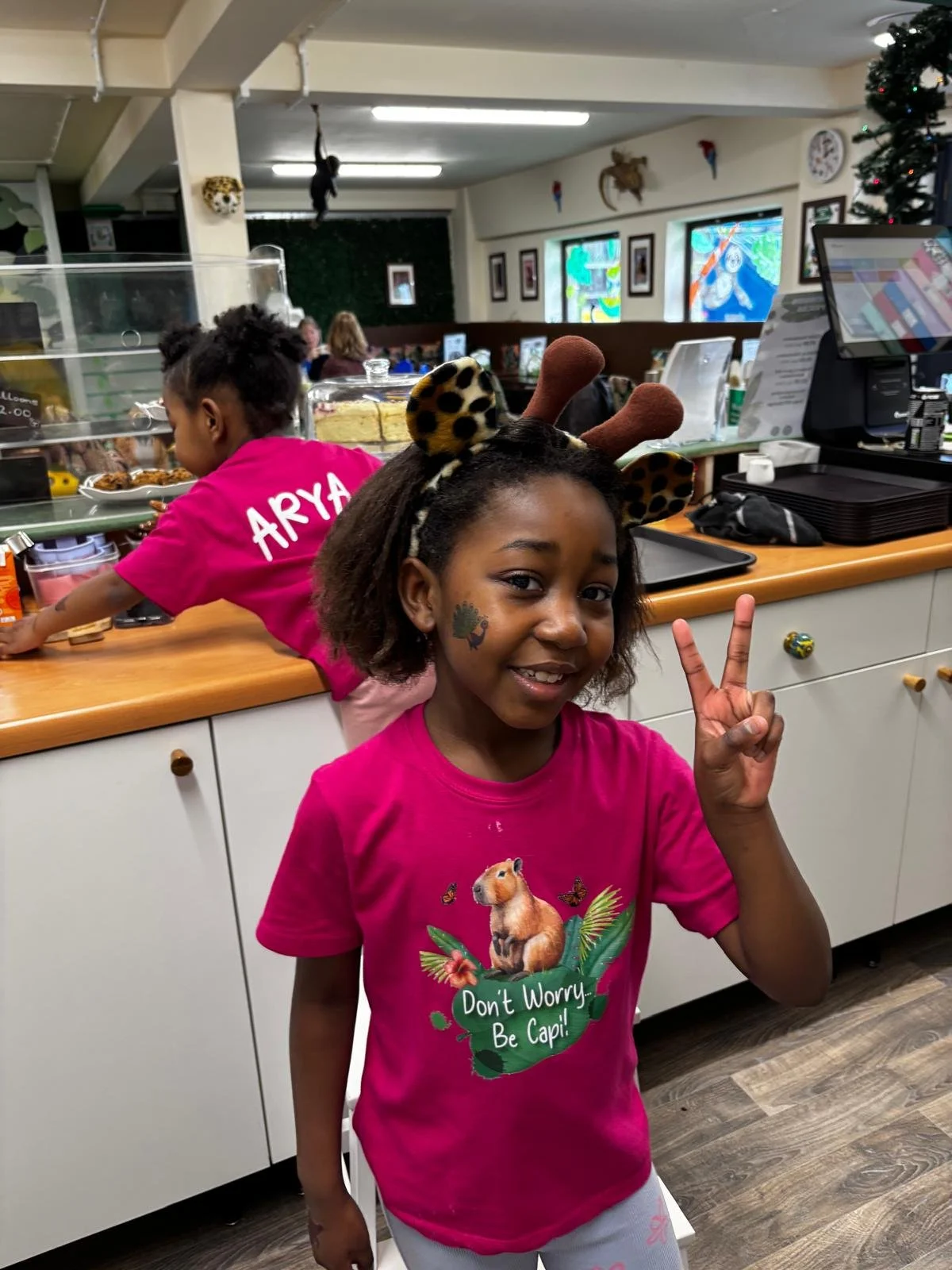 A young girl with a festive reindeer headband making a peace sign inside a cafe, with another girl in a pink shirt behind the counter.