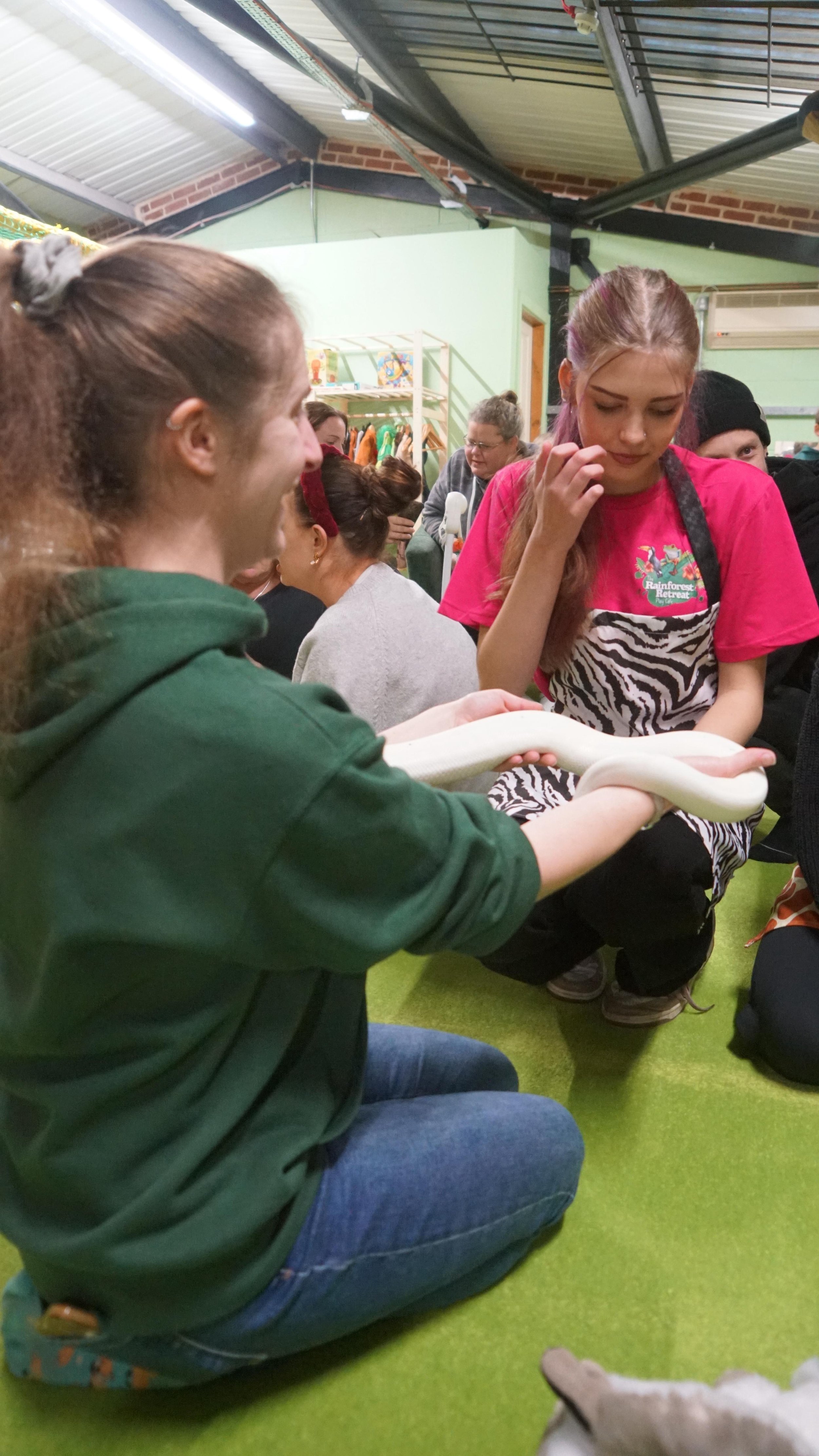 A young woman examining a white snake with a woman sitting on her knees on a green carpeted floor, while other people observe in the background at an indoor event.