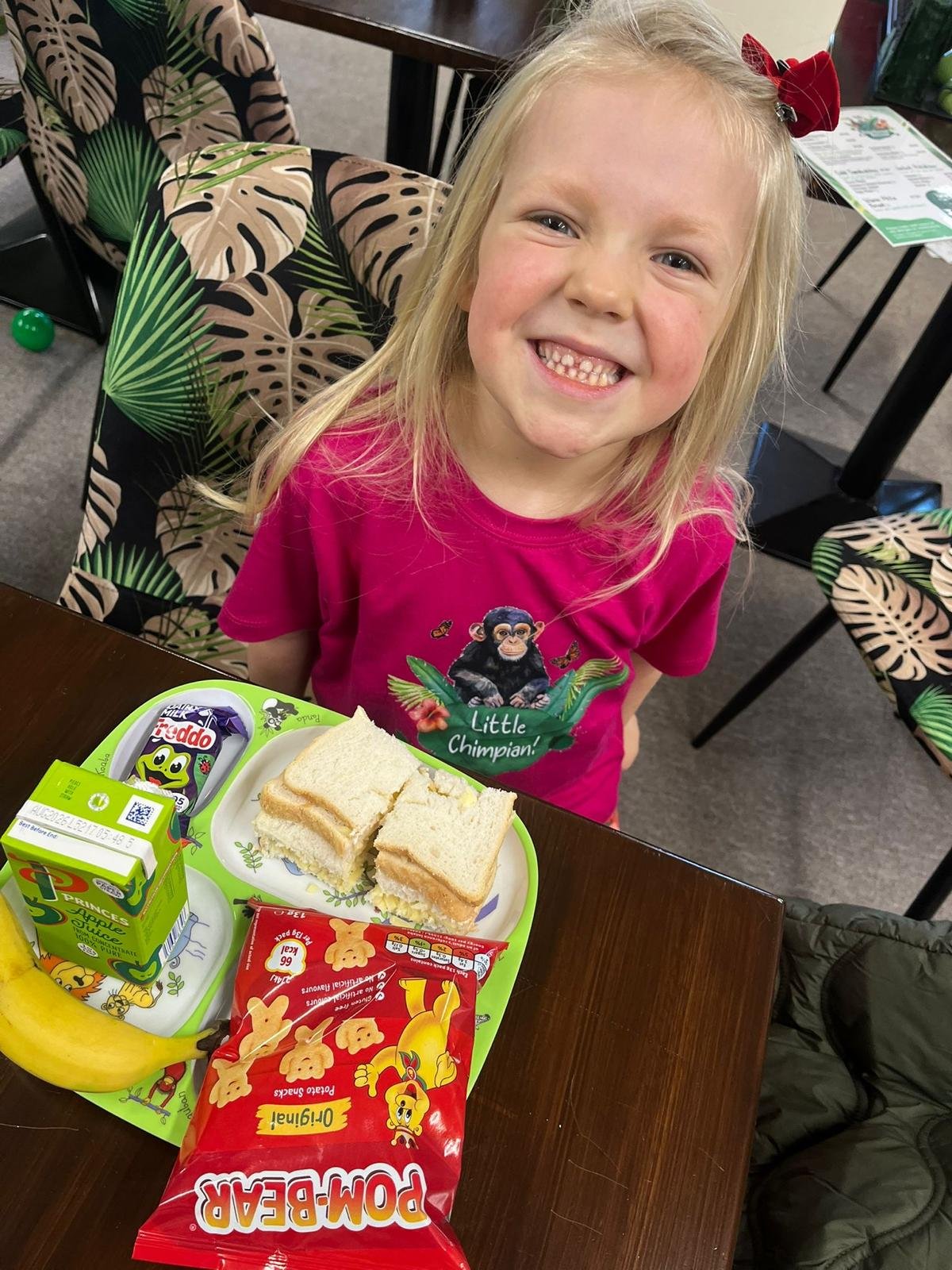 A smiling girl in a pink shirt with a chimpanzee on it sitting at a table with a tray of snacks that include a banana, apple juice, a sandwich, and a bag of animal cookies, in a room with tropical-patterned chairs.