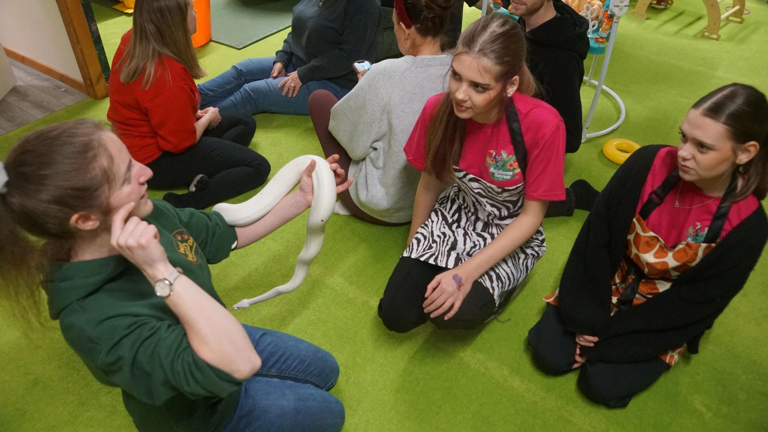 A group of young women sitting on a bright green floor, with two of them kneeling and one of them holding a white snake. The women are engaging in a snake-handling activity, with some watching and others touching the snake. There are toys and equipme