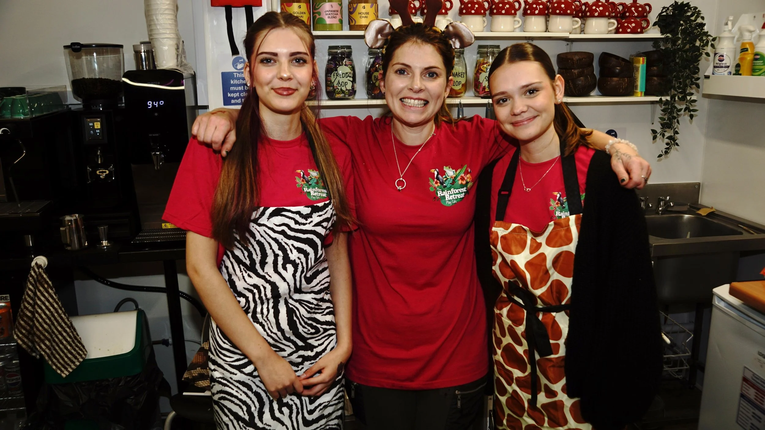 Three women in kitchen, smiling, wearing matching red shirts with a logo, one with zebra stripe apron and another with giraffe print apron, standing close together with arms around each other.