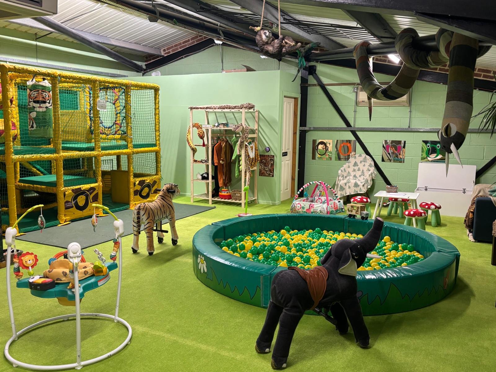 Indoor children's play area with a ball pit, stuffed animal toys, a climbing structure with a tiger plush, small tables and stools, and decorative animal-themed art on the walls.