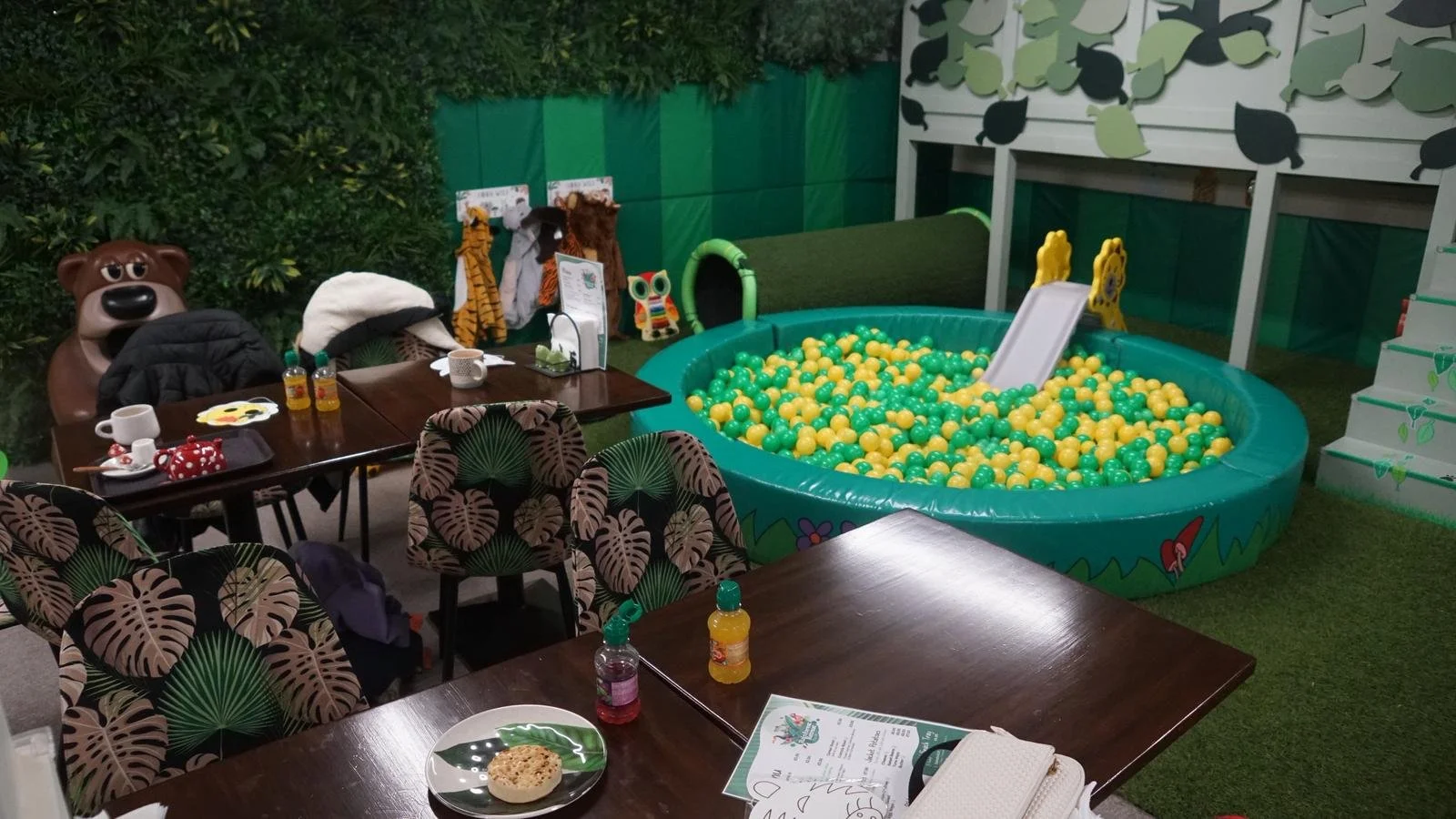 Children's indoor play area with a ball pit, slide, stuffed animals, and tables with children’s drinks and snacks.