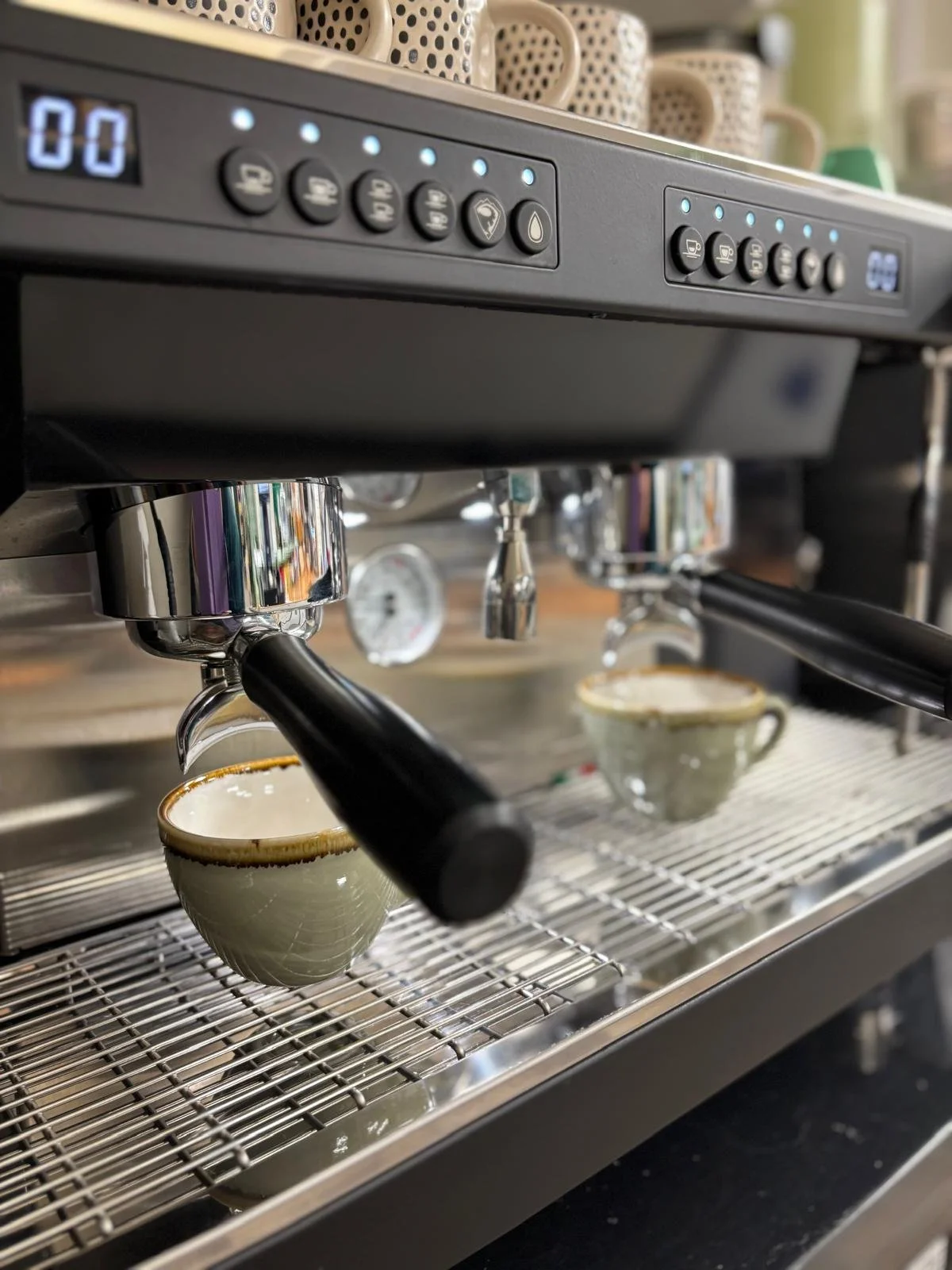 Close-up of a commercial espresso machine brewing two cups of coffee into green ceramic cups with brown rims.