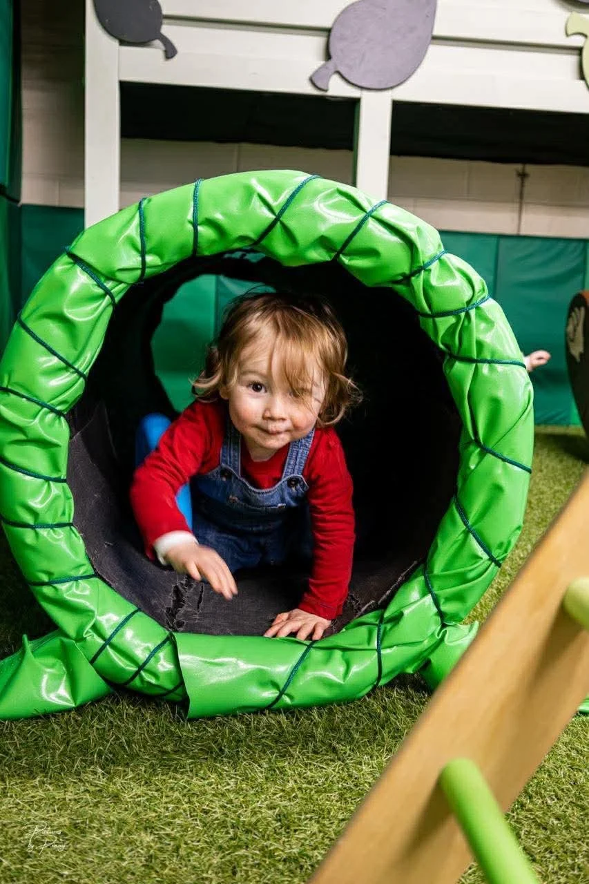 A young child with blond hair, wearing a red shirt and denim overalls, crawling through a green tunnel on a play area with grass flooring.