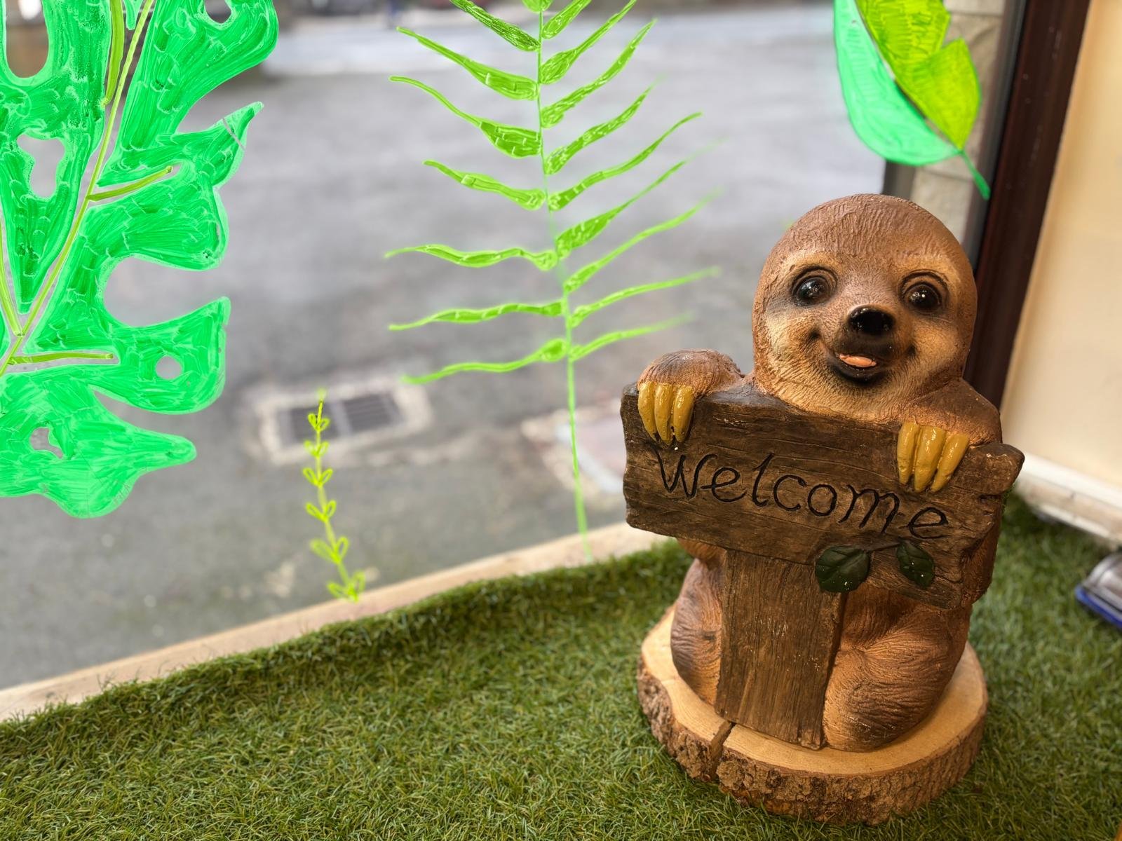A decorative wooden and resin figurine of a baby otter holding a 'Welcome' sign, placed on a grassy surface near a window with painted green leaves on the glass.