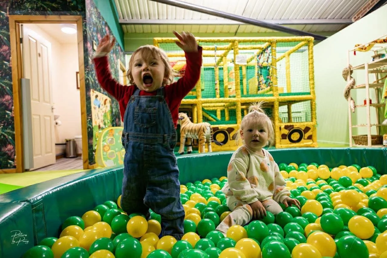Two young children playing in a ball pit at an indoor play area. One child is standing and yelling with arms raised, while the other child is sitting among yellow and green balls. In the background, there is a yellow cage-like play structure with a t