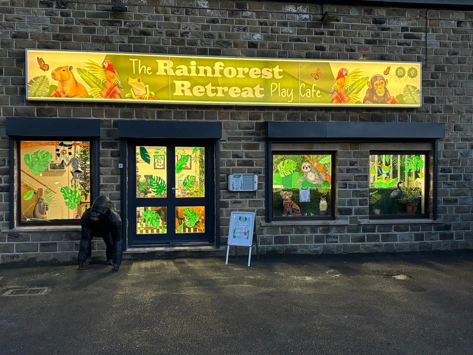 Front of The Rainforest Retreat Play Café building with colorful jungle and rainforest animal decorations on windows, including a gorilla statue outside, and a bright sign with jungle animals at the top.