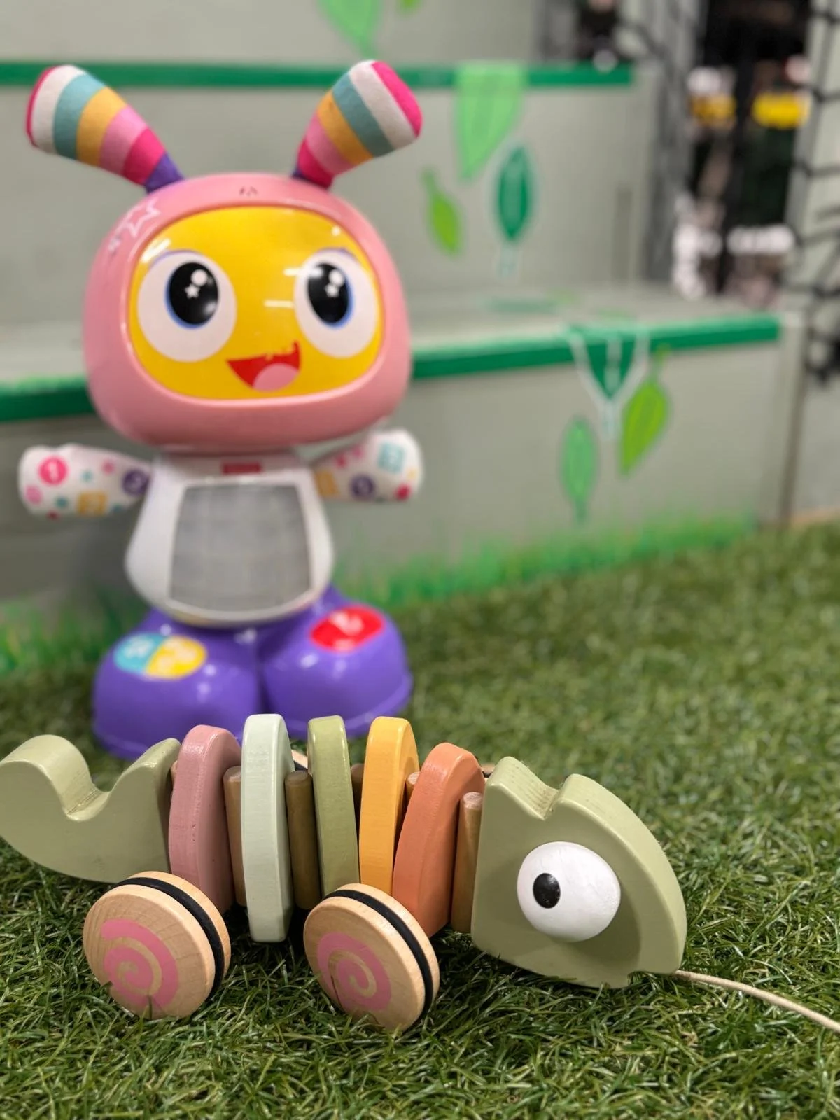 Colorful wooden toy chameleon with large eyes, placed on artificial green grass, with a colorful cartoon robot with a smiling face and antennae in the background.