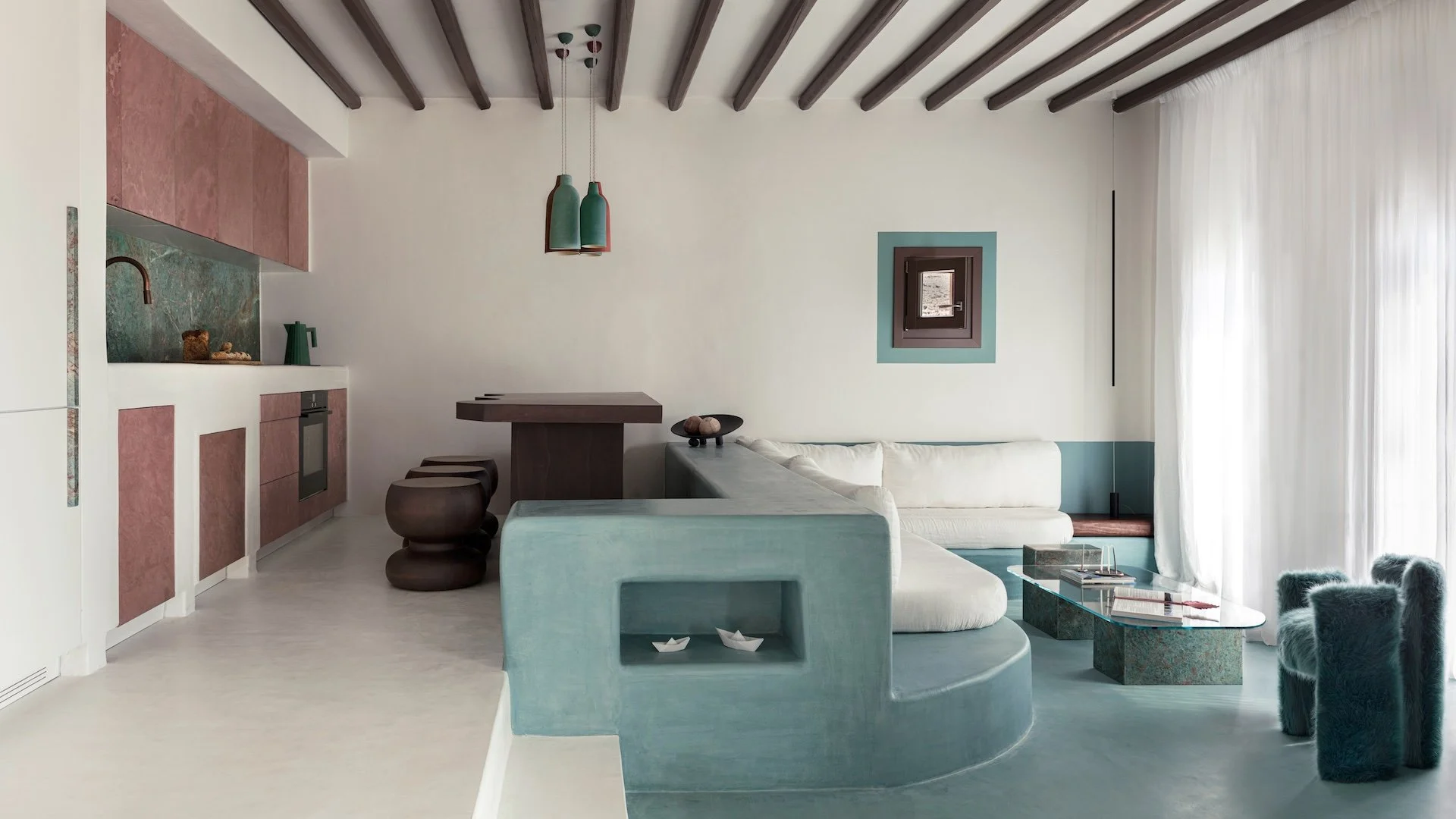 Modern living room with a white couch, blue and green accents, a small window, and a kitchen area with pink cabinets and a green backsplash.