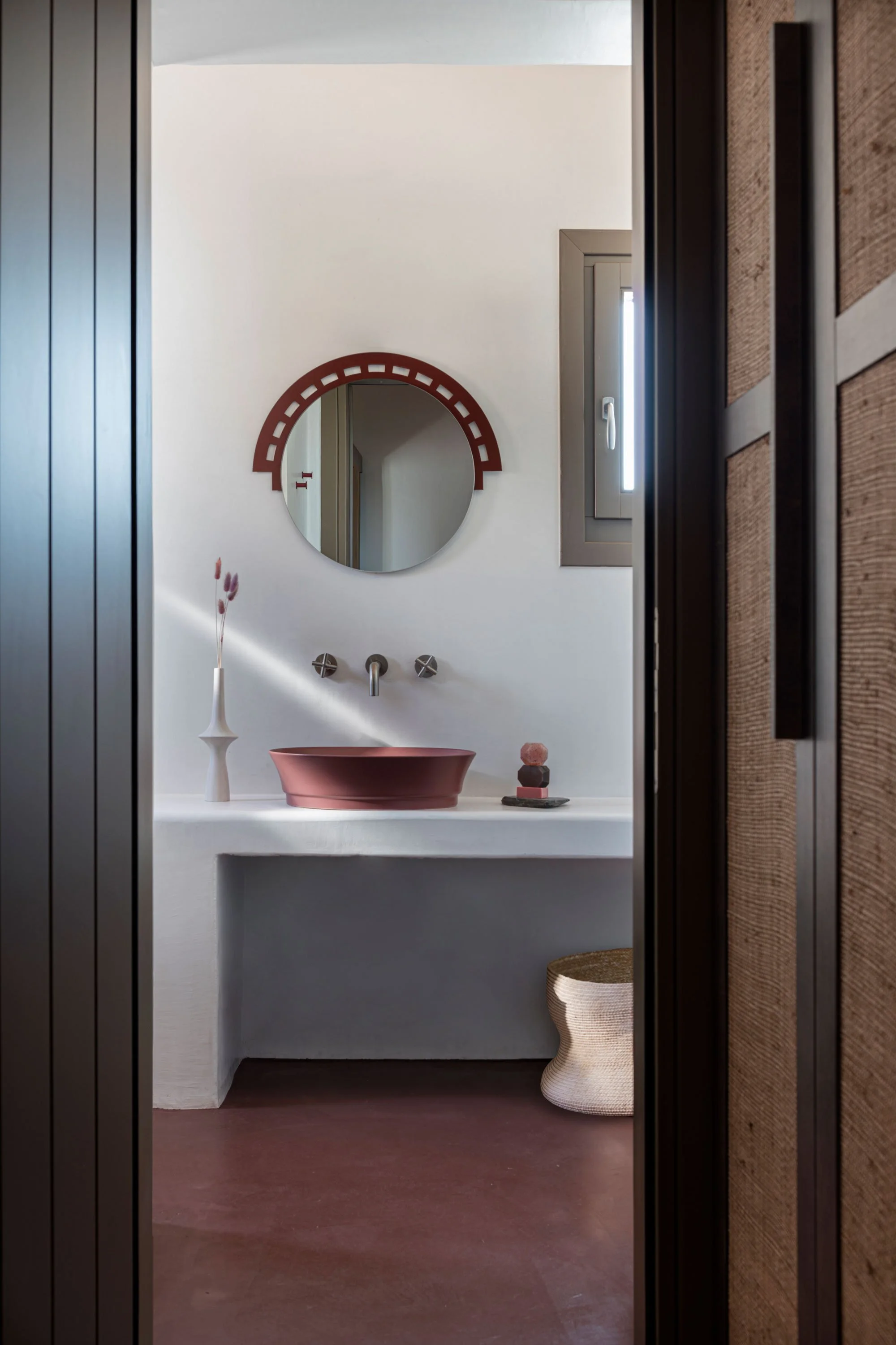 Interior view of a minimalist bathroom with a pink vessel sink, a round mirror with a decorative frame, a small window, a vase with dried flowers, and a woven basket, seen through a doorway with dark wooden framing.