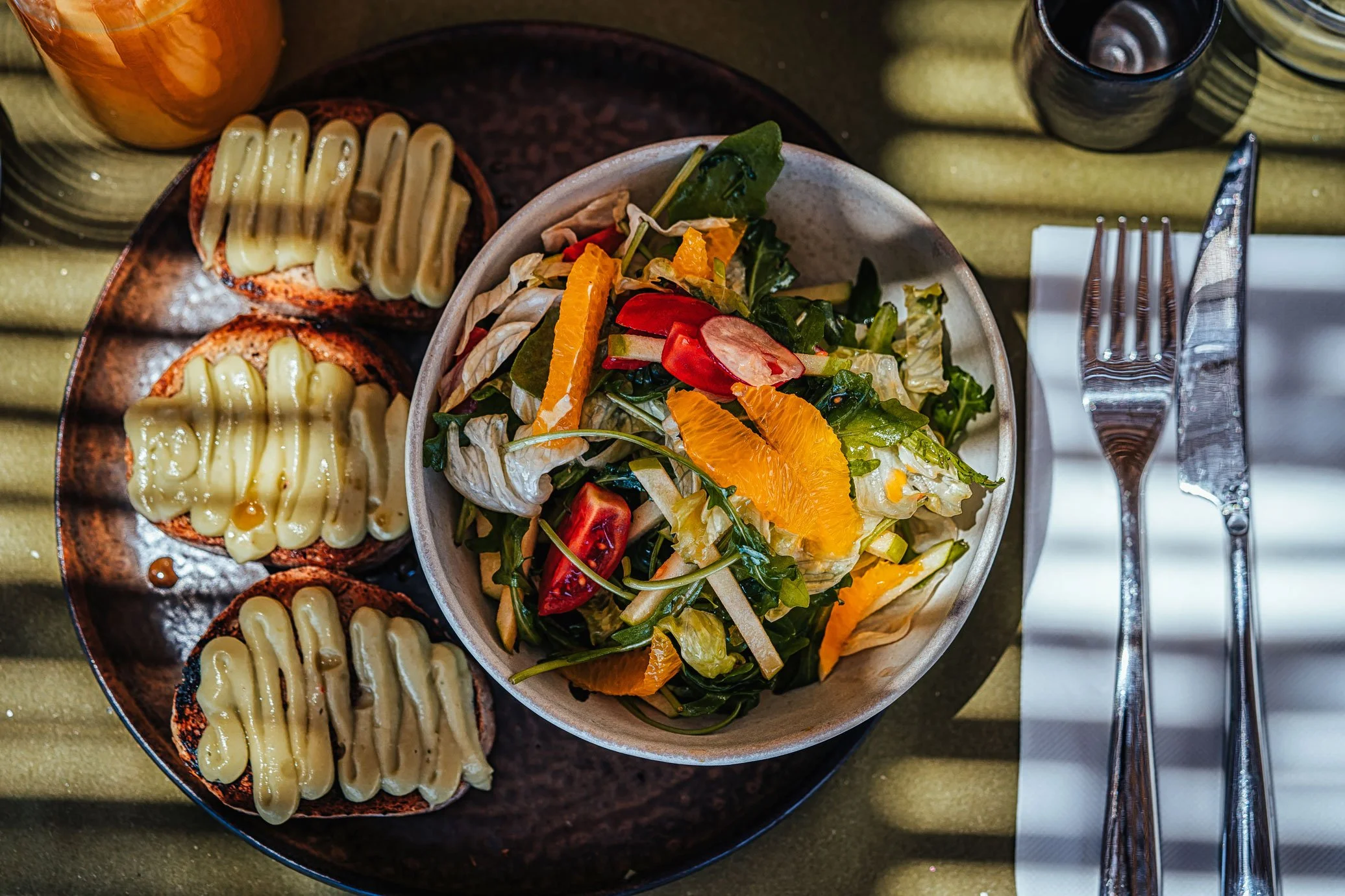 A bowl of mixed salad with orange slices and cherry tomatoes, served alongside three slices of bread with melted cheese, on a dark plate. Fork and knife are beside the bowl on a white napkin.