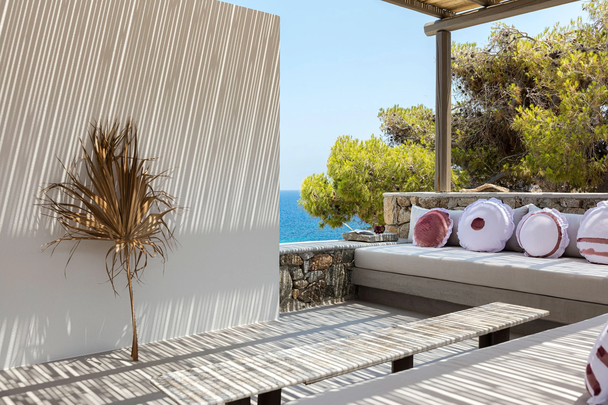 An outdoor seating area with a cushioned bench and decorative pillows overlooking the ocean, framed by lush green trees, with sunlight casting striped shadows.