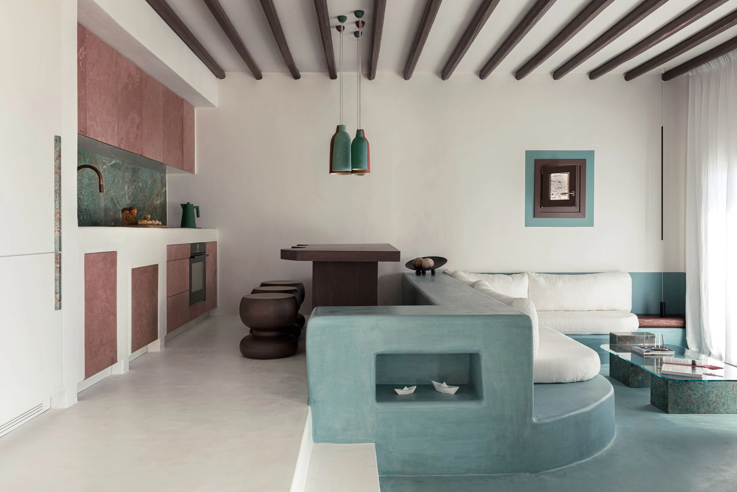 Living room with white walls, dark wooden ceiling beams, and a small window. There is a white sectional sofa with a blue base, a coffee table with a marble look, and a small restaurant-style table with stools. The room features minimalist decor with 