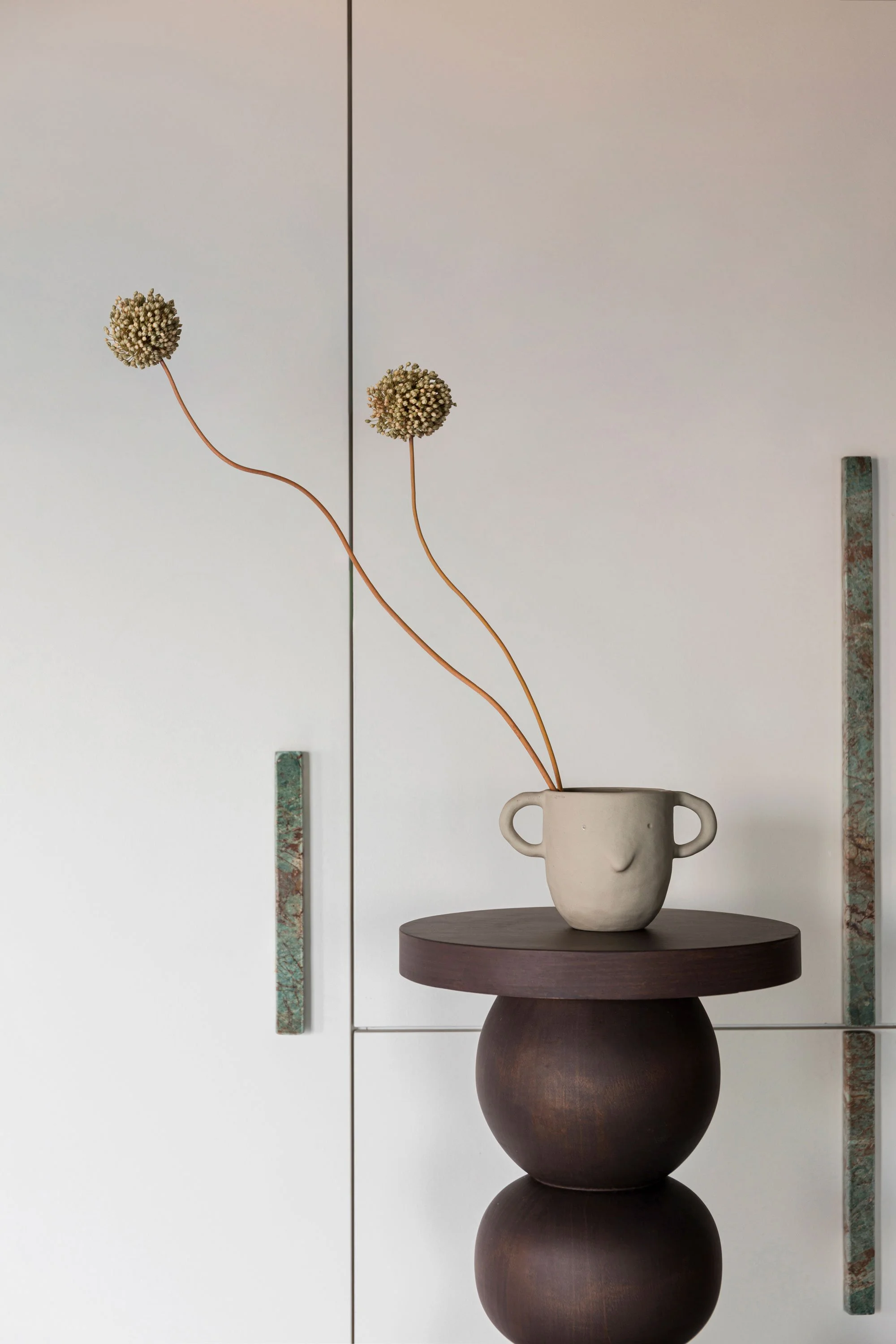 A minimalist ceramic face vase with two handles containing two dried flower stems, placed on a round dark wooden table with a modern white cabinet in the background.