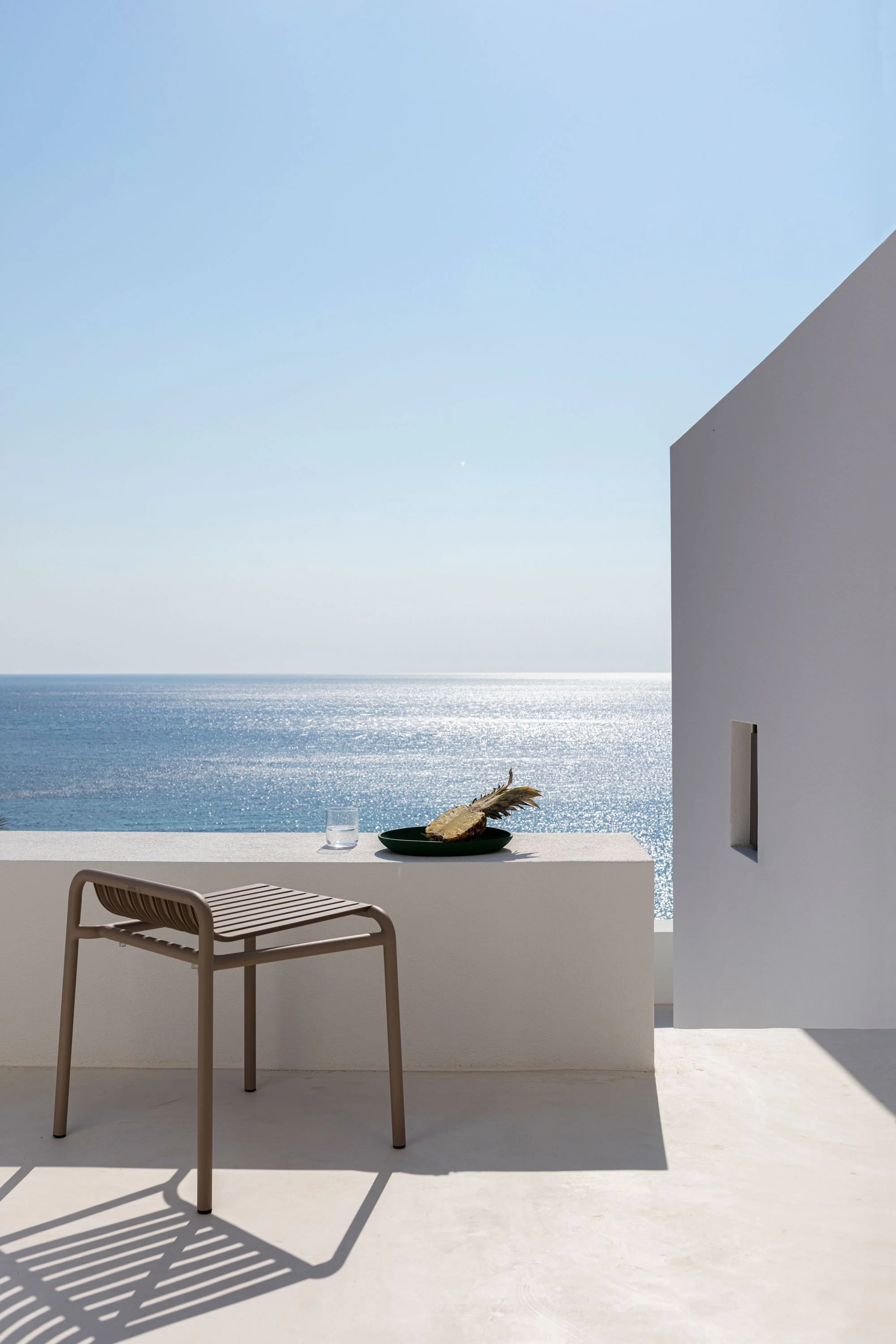 Modern outdoor terrace with a beige chair, a black bowl with bread and a leaf, a glass of water, white walls, and ocean view under a clear blue sky.