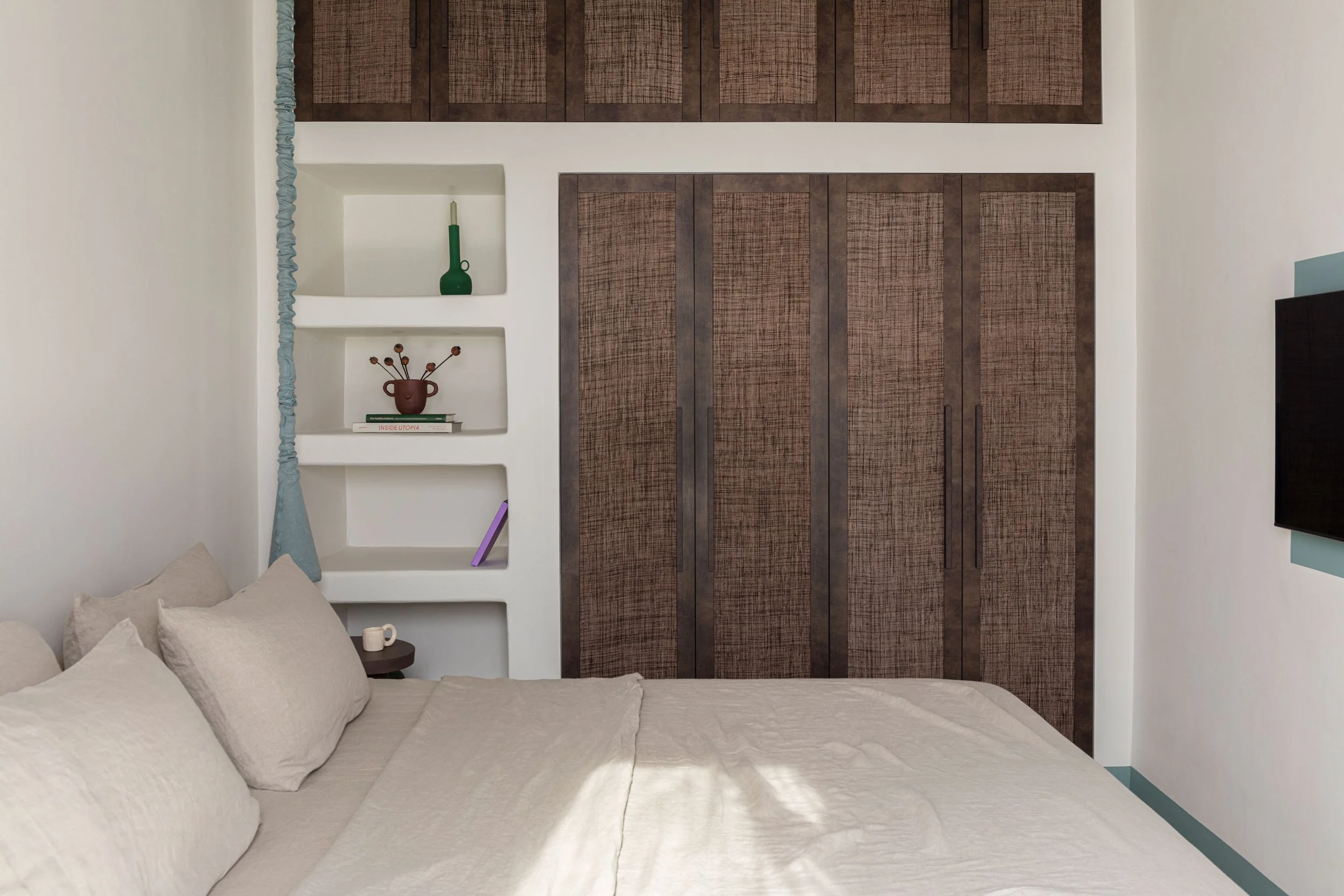 A bedroom with a bed covered in beige bedding, a small side table with a cup, a built-in bookshelf with decorative items, and a large brown closet with textured doors.