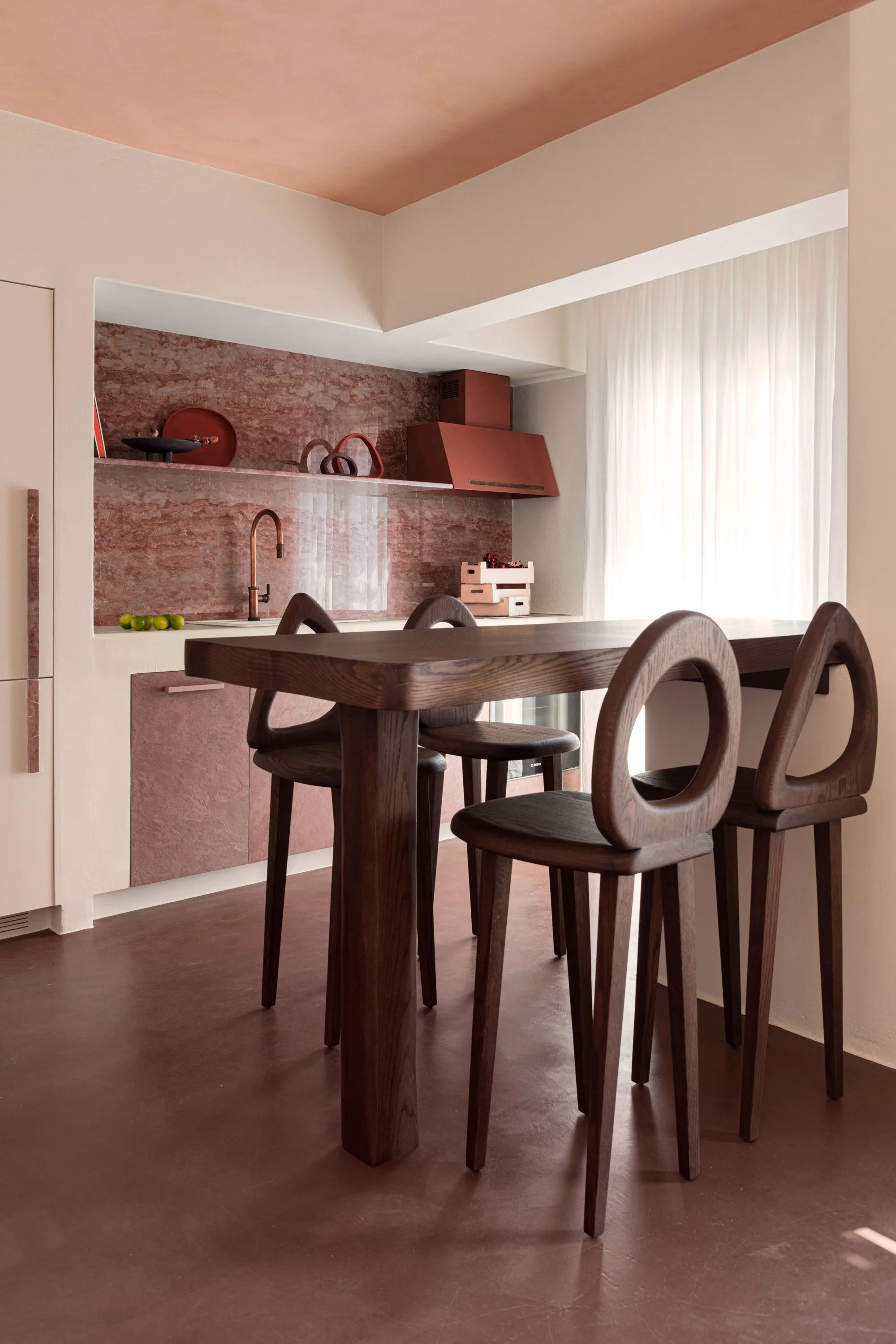 A dining area with a dark wooden table and three matching wooden chairs in front of a pink-themed kitchen with a brick backsplash, pink cabinets, and a window with white curtains.