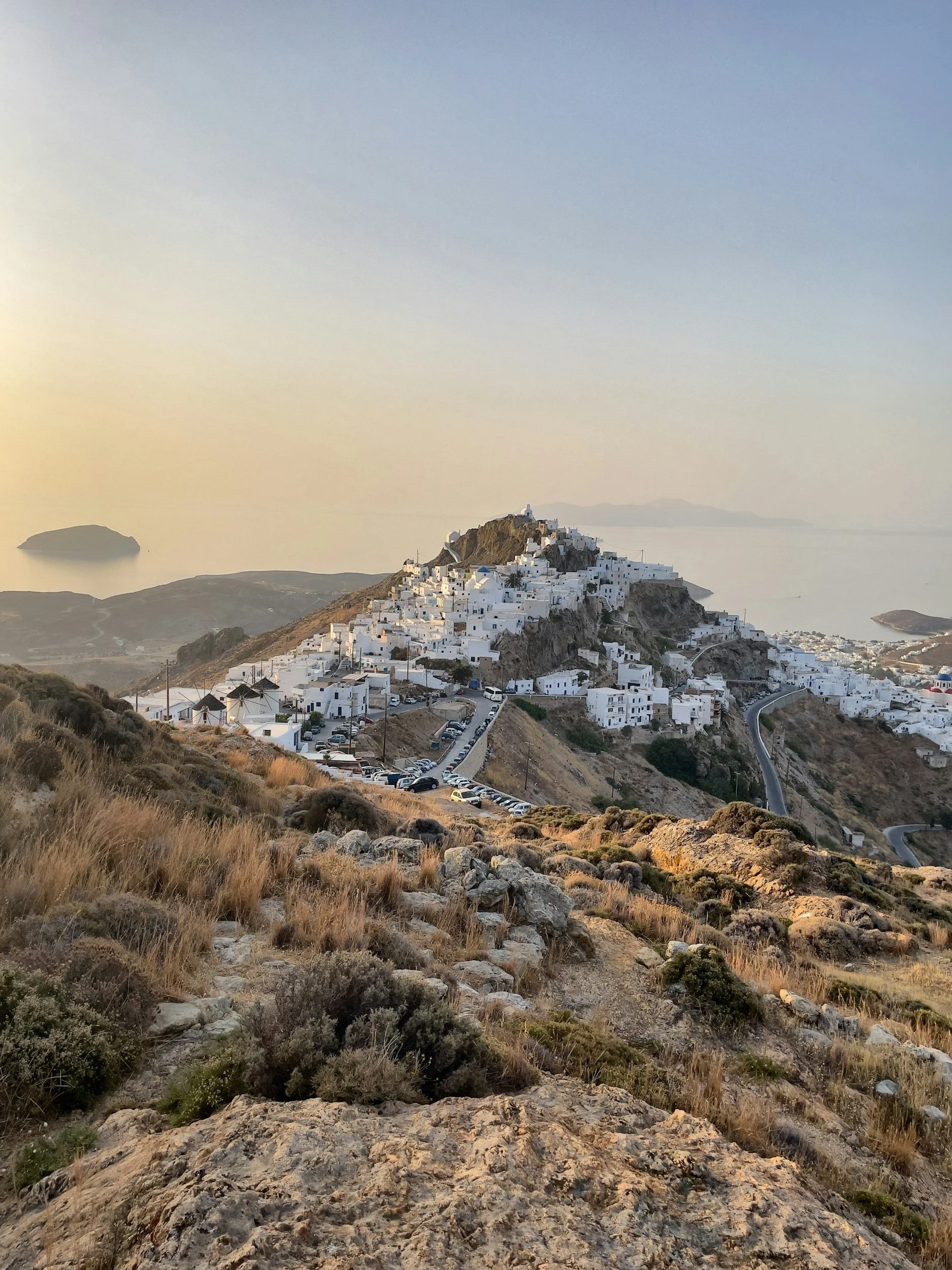 A scenic view of a hillside town with white buildings, narrow winding roads, and a parking lot in the foreground, overlooking the sea at sunset.