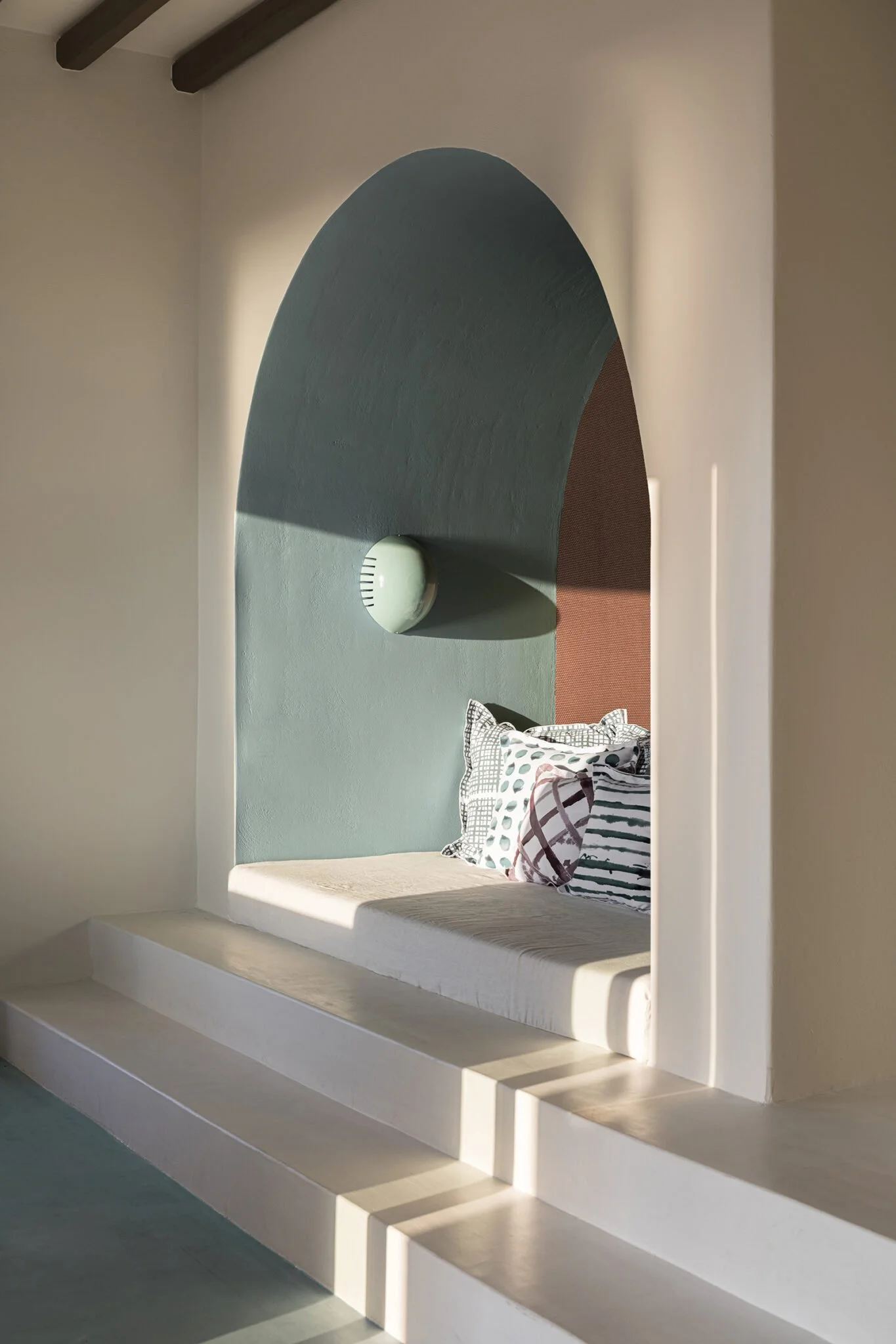 A built-in bed nook with a light blue wall accent, three striped and patterned pillows, surrounded by a white plastered arch and steps, with a modern wall sconce.