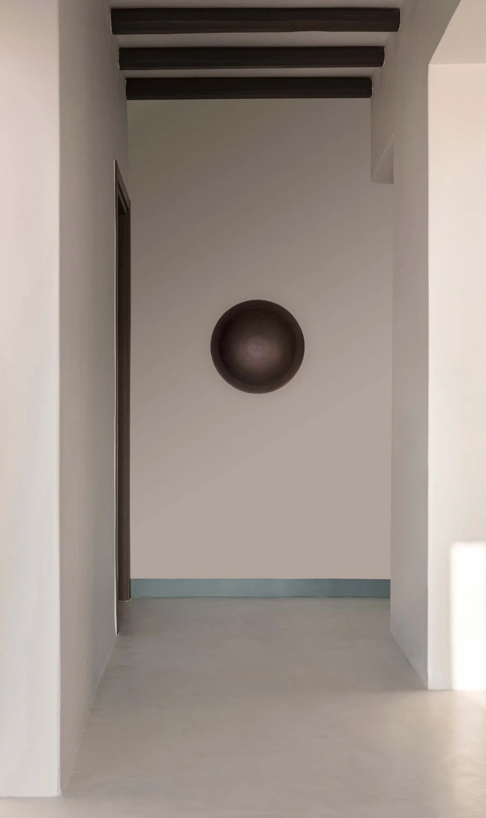 Empty hallway with white walls, a round black decorative object on the wall, and dark wooden ceiling beams.
