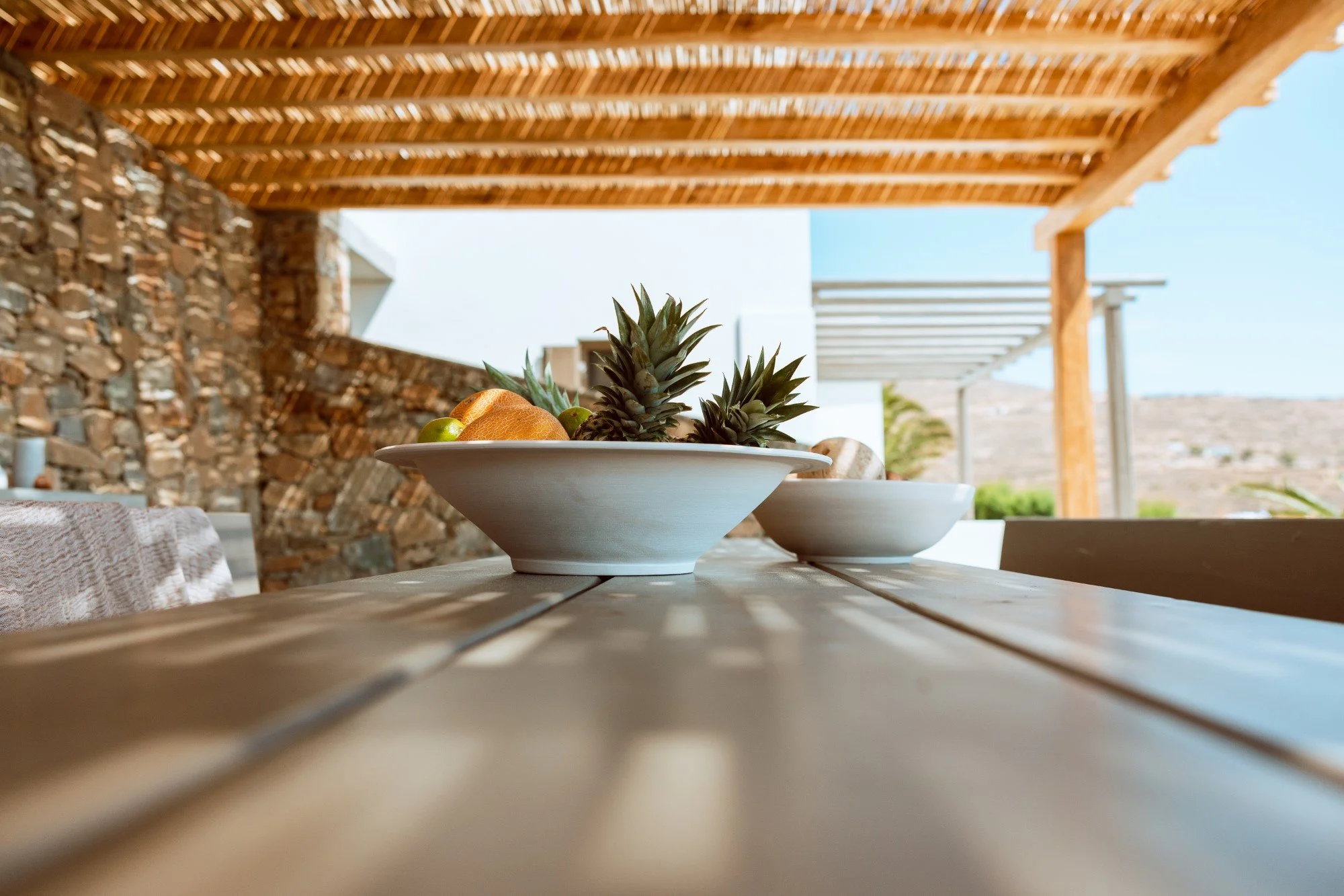A wooden outdoor table with white bowls filled with fruits, including a pineapple, grapes, and an orange, under a wooden pergola.