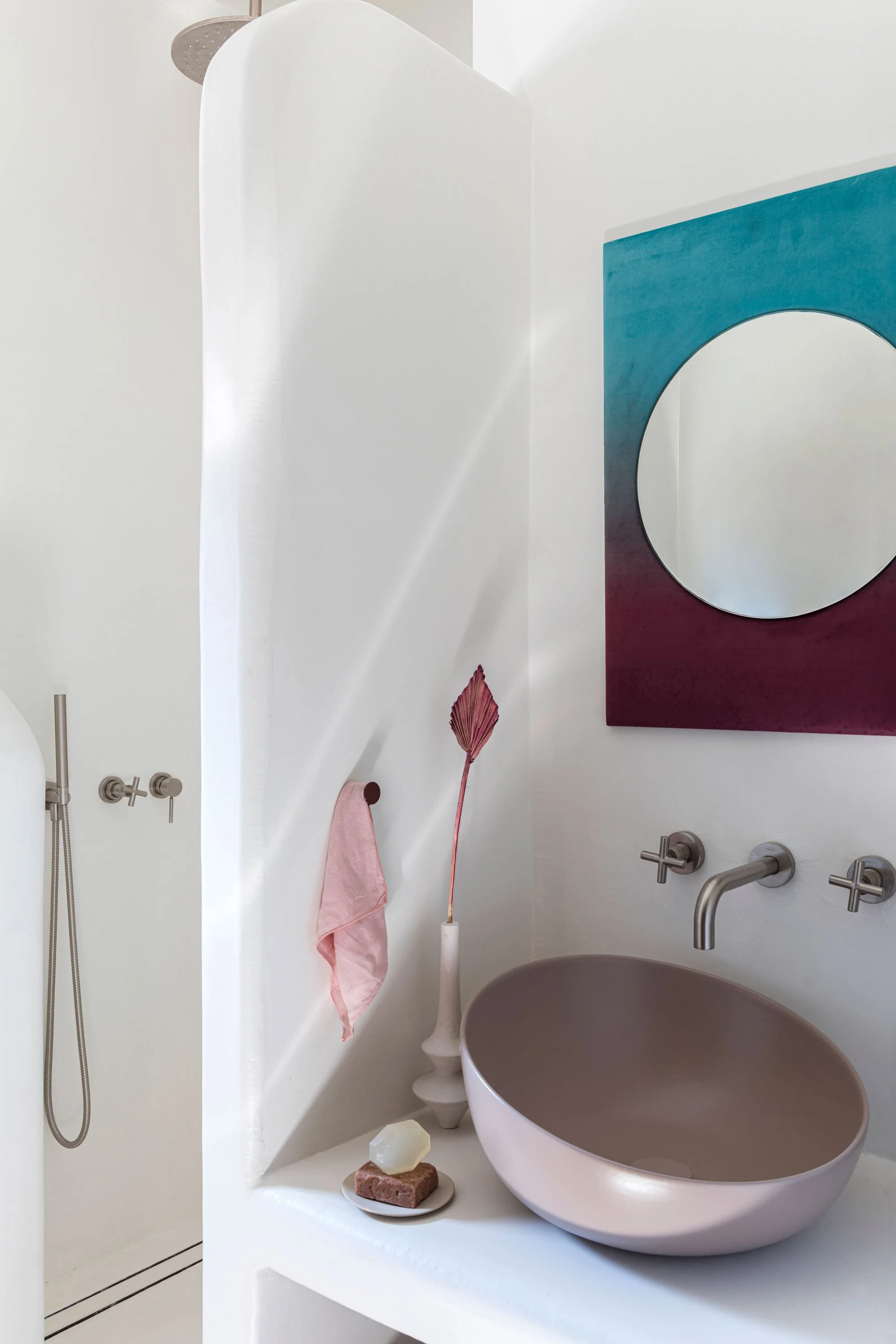 Modern bathroom with a round beige vessel sink, wall-mounted faucet, pink towel, abstract blue and purple wall art, decorative vase with a dried leaf, soap on a dish, and a walk-in shower with a handheld showerhead.