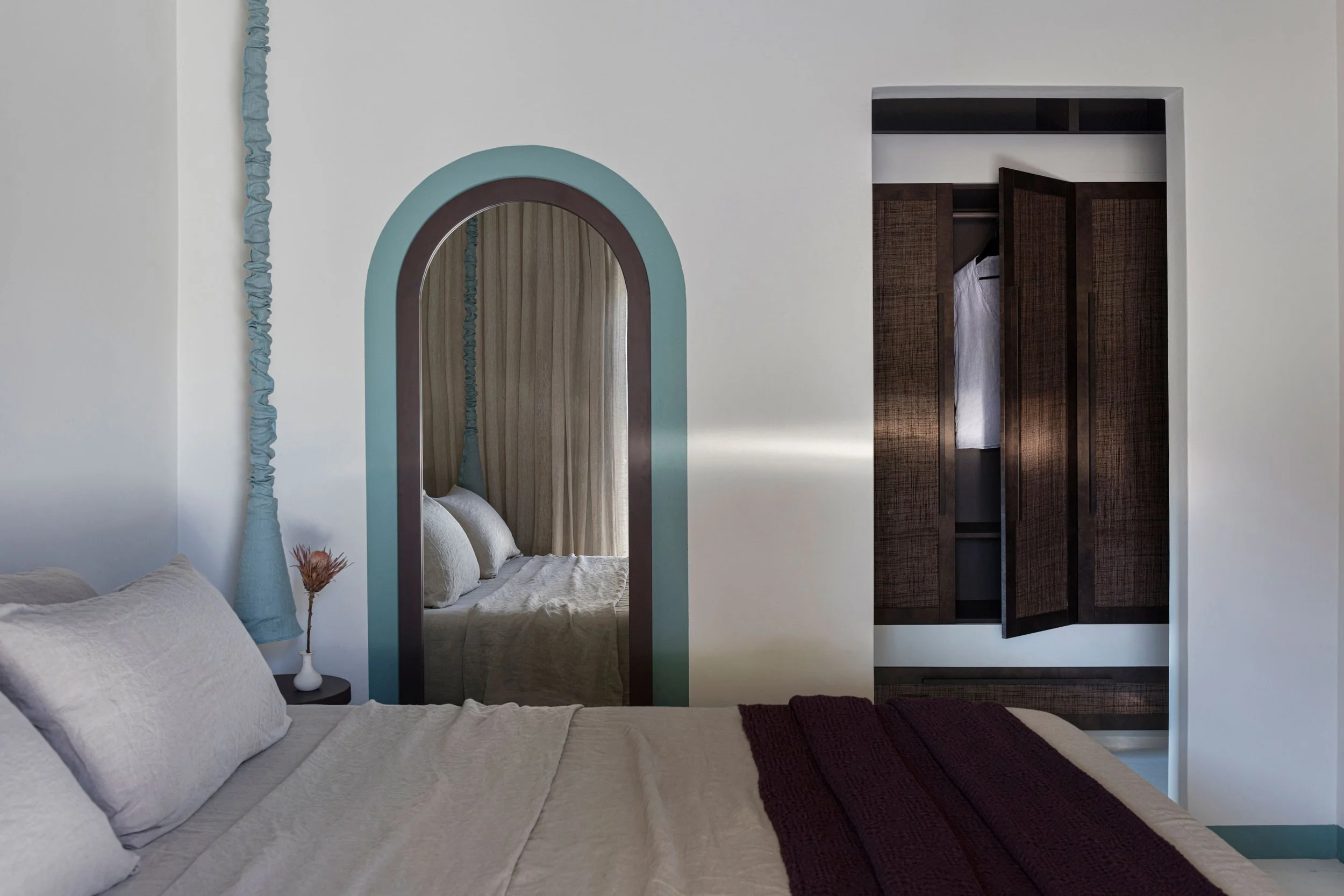 A bedroom with a bed covered in beige linens, a small bedside table with a vase and dried flower, a decorative mirror with a blue and brown arch design reflecting the bed, and a closet with dark wooden doors, one of which is slightly open.