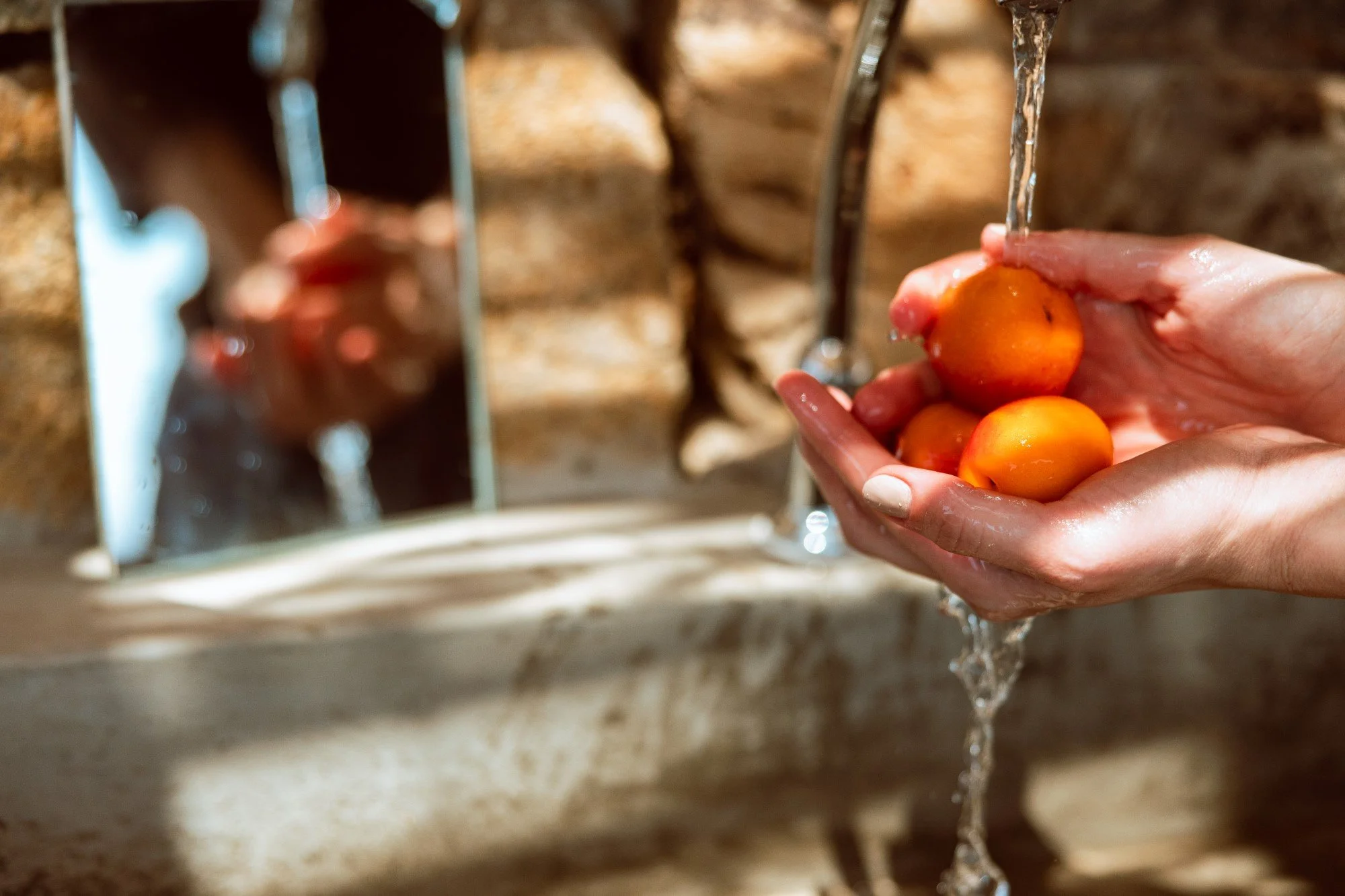 A person washing small orange tomatoes under running water in their hands at a sink.