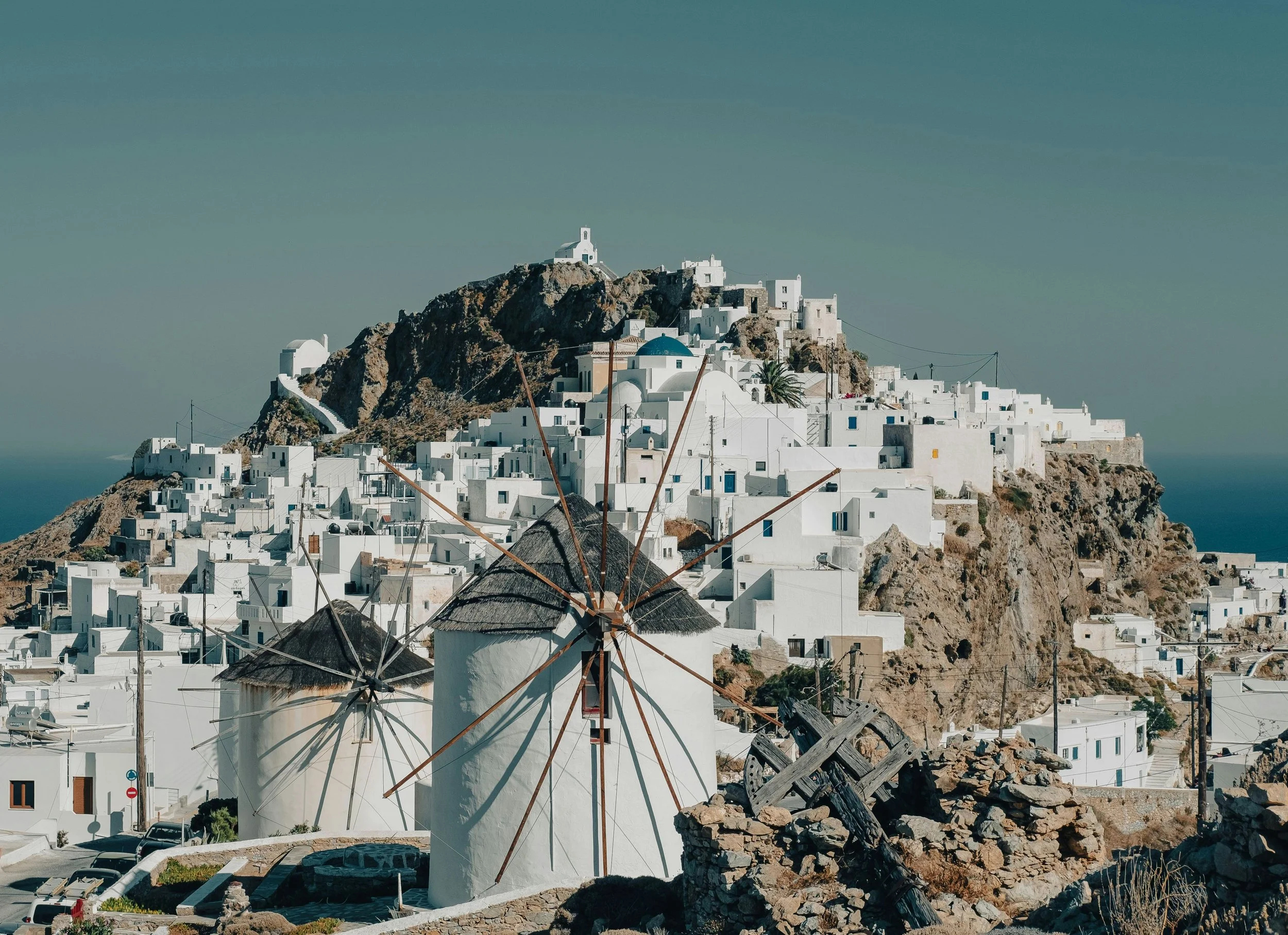 A scenic view of a white-washed village on a hillside with windmills in the foreground, characteristic of Santorini, Greece, under clear blue skies.