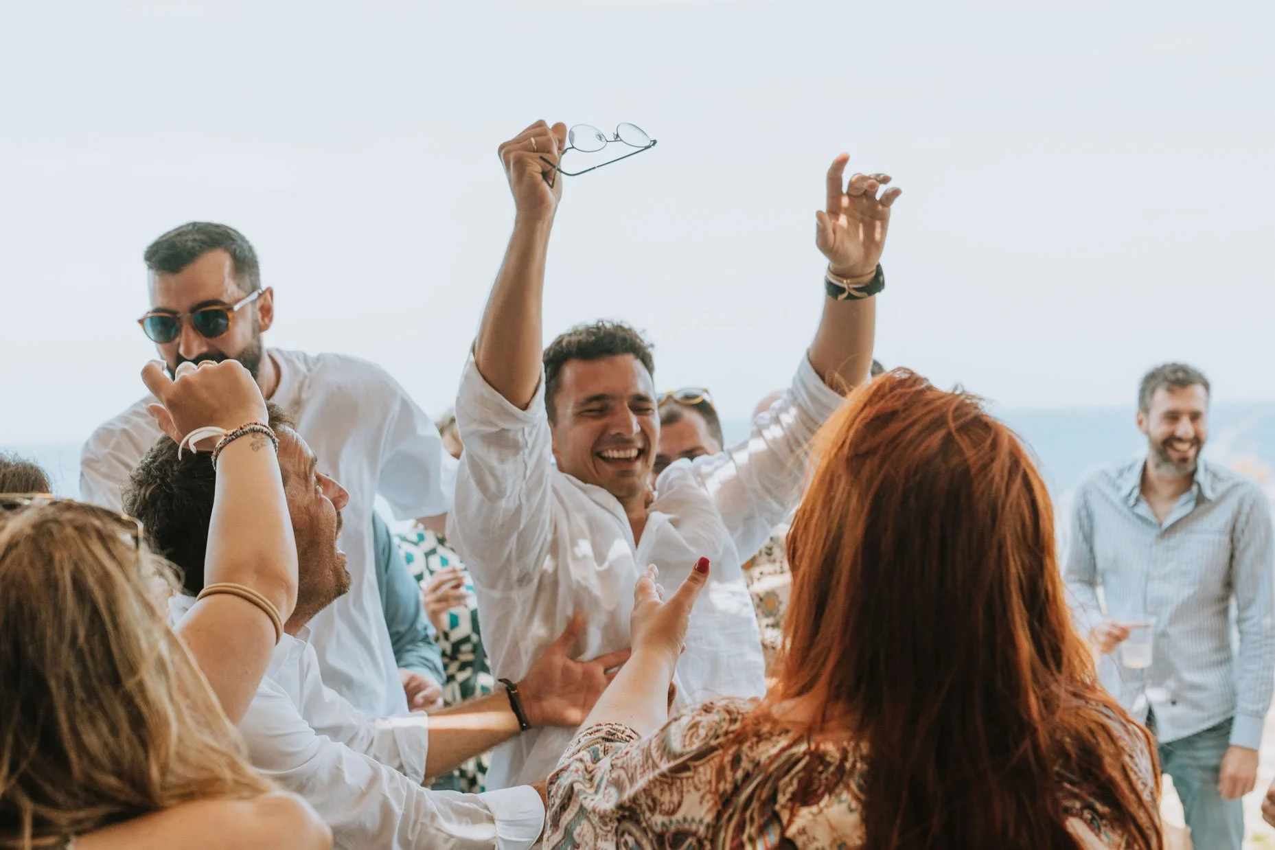 People enjoying a sunny outdoor celebration, smiling and dancing, some wearing glasses and casual clothes.