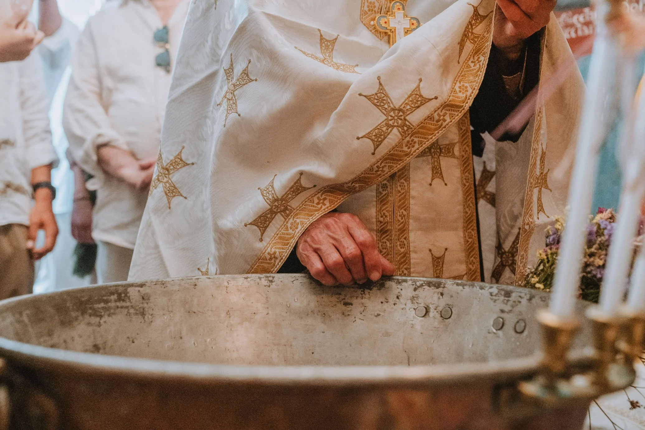 A priest wearing ornate gold-embroidered white robes performs a baptism ritual, with hands near a baptismal font.