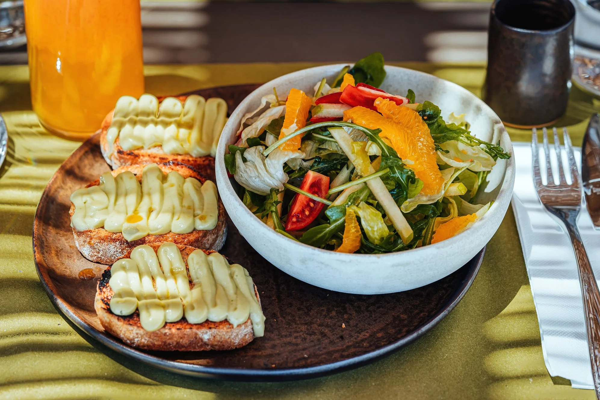 A plate with three slices of toasted bread topped with melted cheese, placed next to a bowl of mixed salad containing lettuce, cherry tomatoes, orange slices, and other vegetables. A glass of orange juice is partially visible in the background, along