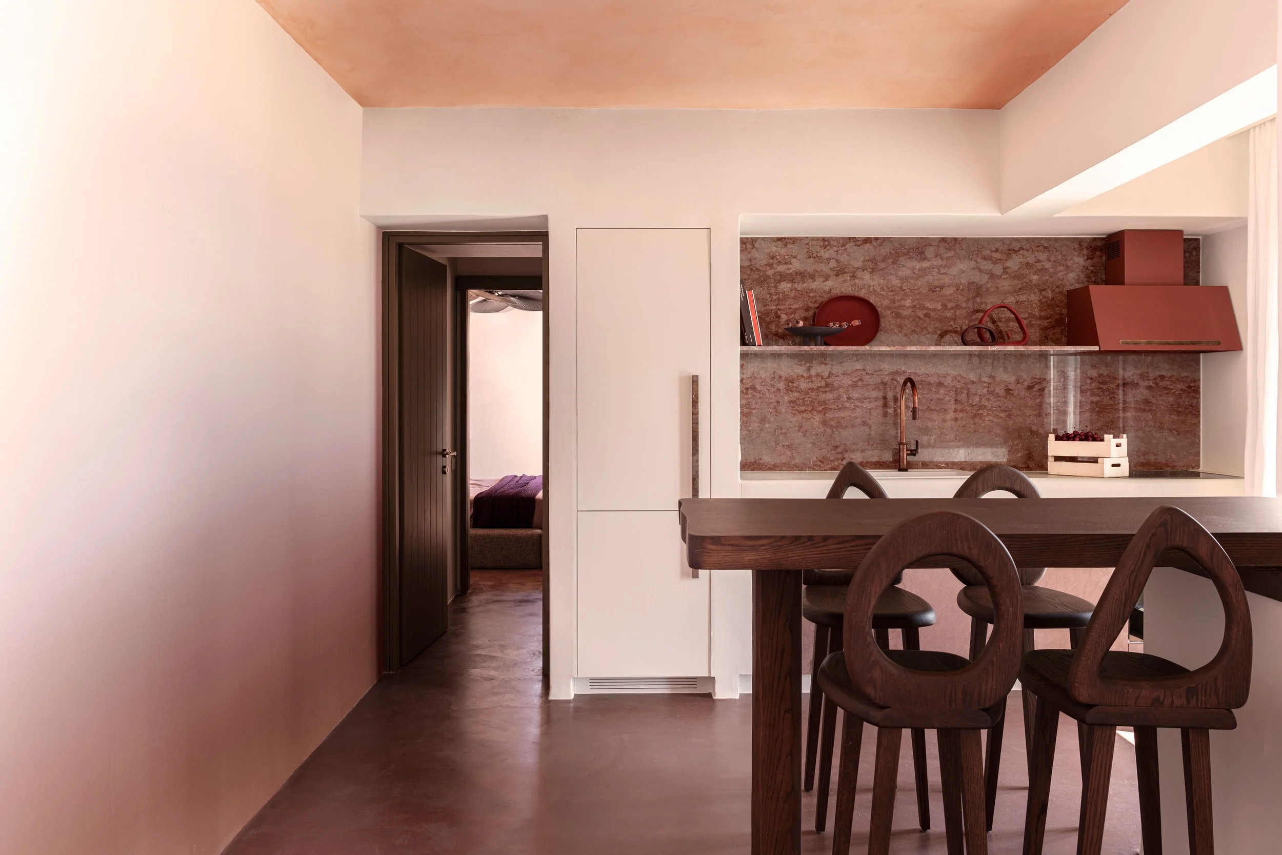 Modern kitchen with wooden dining table and chairs, open to a bedroom with a bed, and doorways leading to other rooms, neutral color palette, textured backsplash in the kitchen.